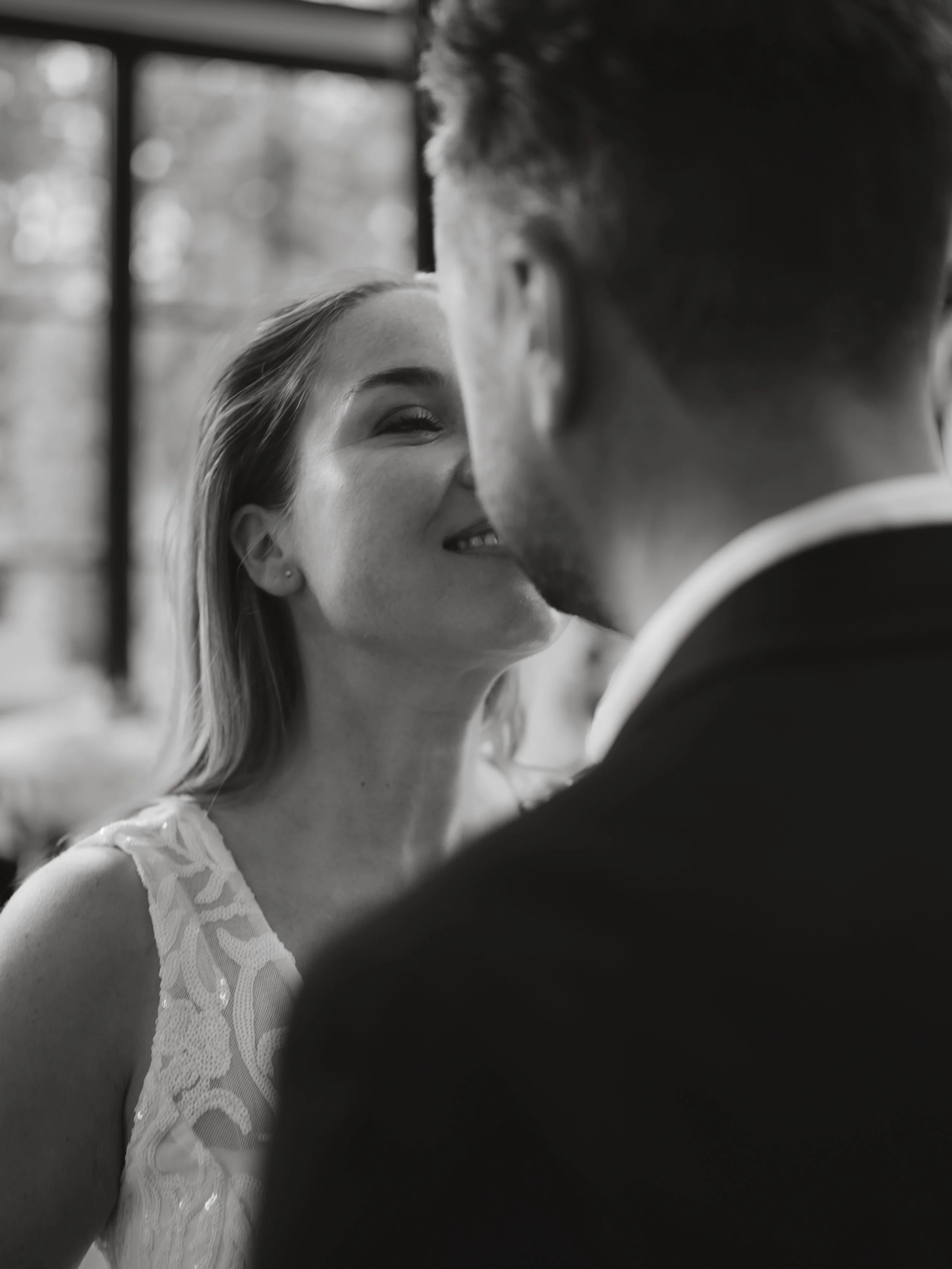 A woman in a lace dress is smiling as she leans in close to a man, possibly during a romantic moment. The photo is in black and white with a blurred background.