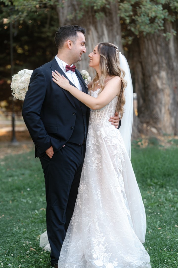 A bride and groom hugging and smiling in an outdoor setting with trees in the background. The groom is wearing a navy suit with a bow tie, and the bride is in a white lace wedding gown with a veil, holding a bouquet of white flowers.