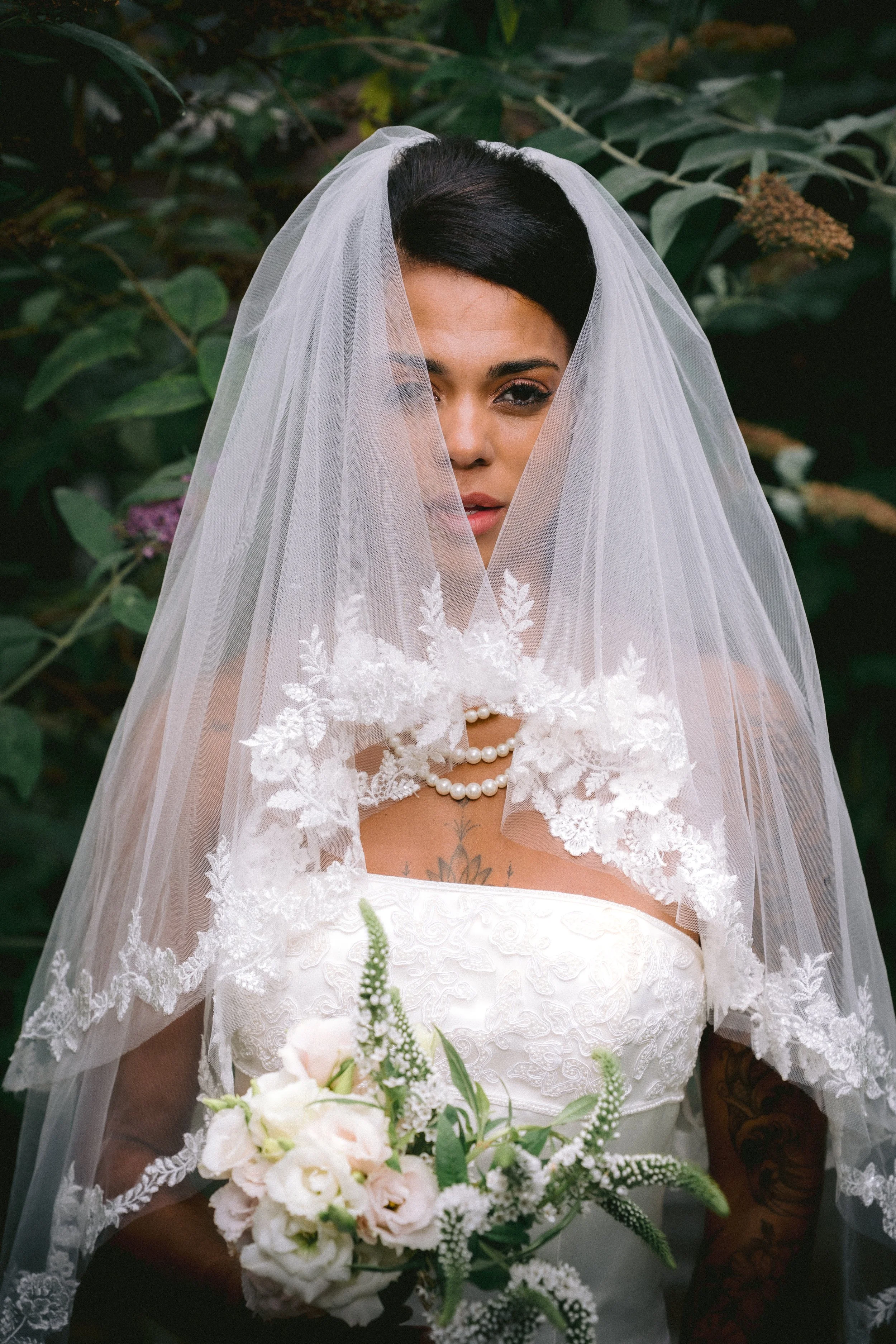A woman dressed as a bride holding a bouquet of white and light pink flowers, with a veil covering her face and shoulders, blue eyes, dark hair, and a lace dress, standing outdoors among green foliage.