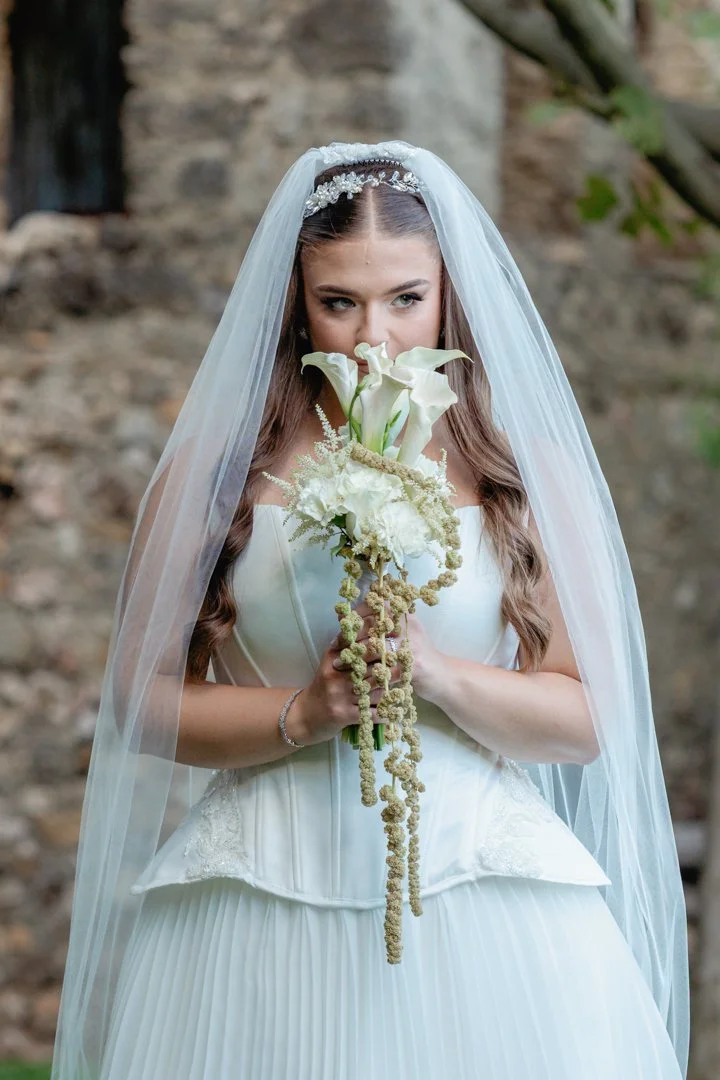 A bride in a white wedding dress and veil holding a bouquet of calla lilies and white flowers outdoors.