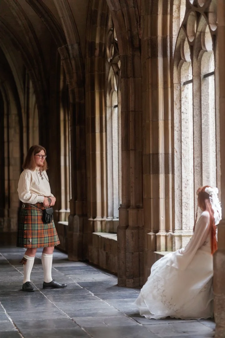 A woman dressed in a Scottish kilt and white blouse stands inside a stone church, taking a photo of a young girl in a white dress who is sitting on a stone bench near tall arched windows.