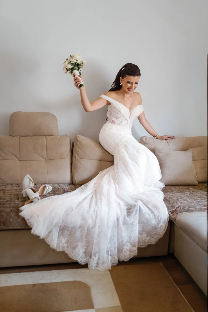 A bride in a white wedding dress sitting on a beige sofa, holding a bouquet of white flowers in her right hand, with her shoes placed beside her on the sofa, smiling happily.