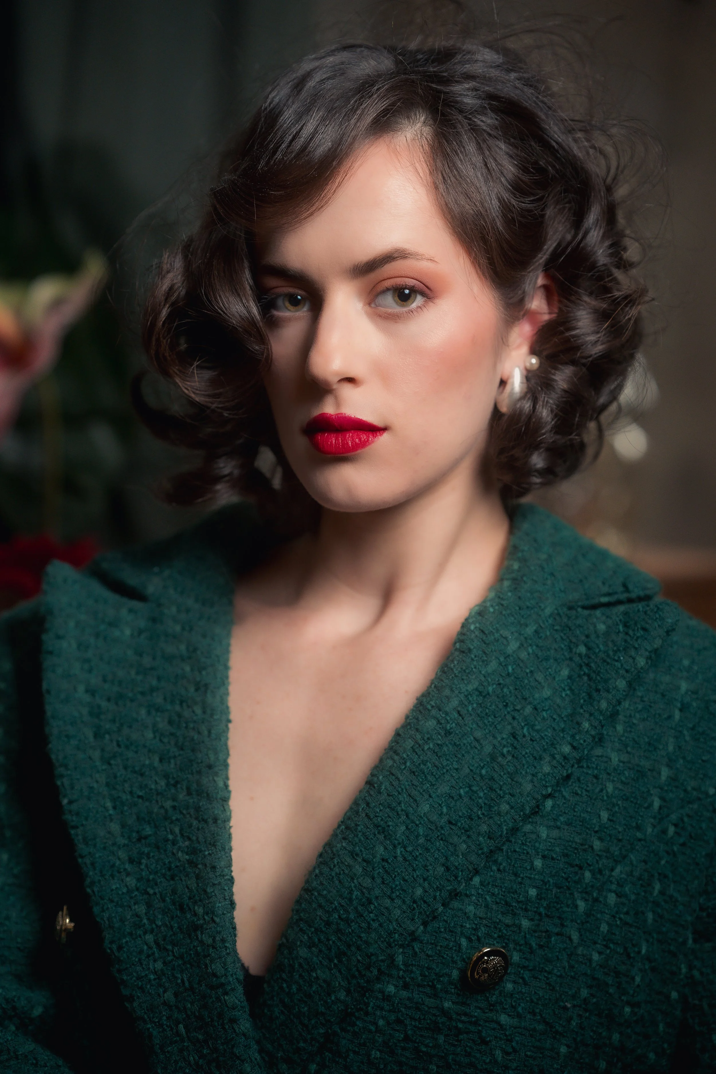 A young woman with dark, curly hair and blue eyes, wearing red lipstick, pearl earrings, and a green textured coat with a black button, standing indoors.