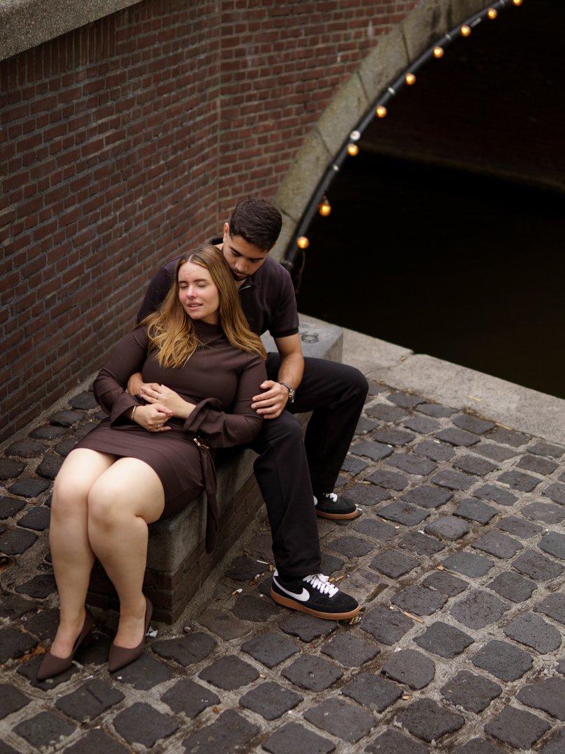 A young couple sitting on a stone ledge beside a brick wall and a canal at night. The woman is wearing a brown dress and heels, and the man is in black pants and sneakers. They are close, with the woman leaning back against the man, who has his arm around her.