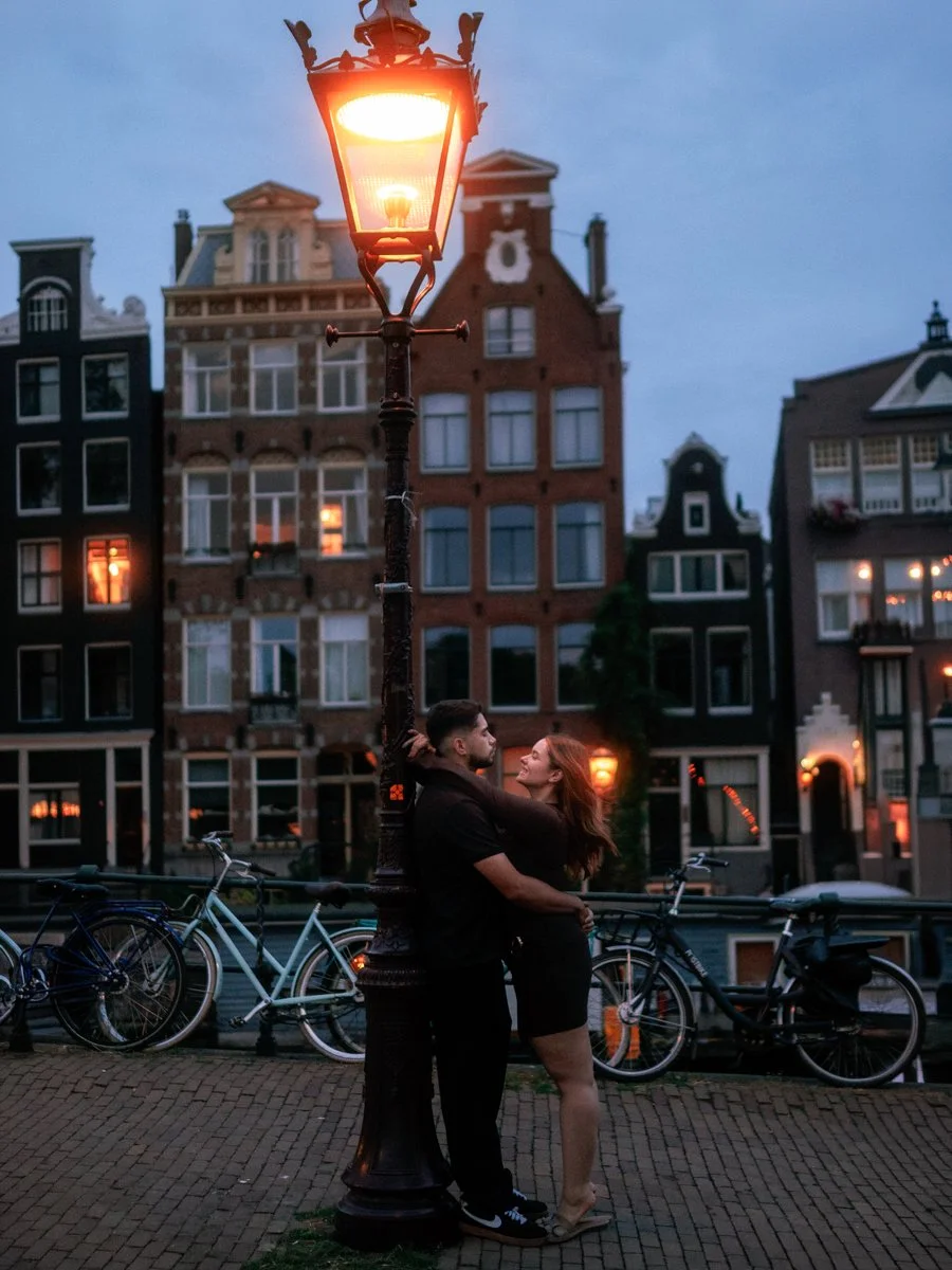 Couple hugging under a streetlamp at dusk, with historic buildings and bicycles in the background.