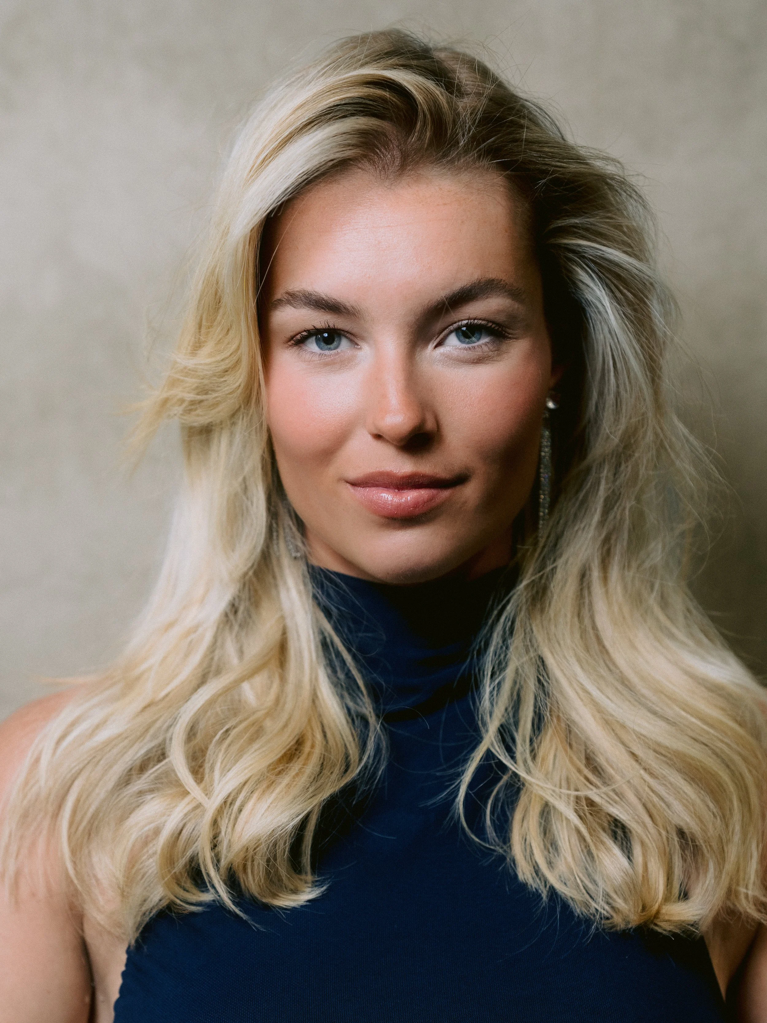 Close-up portrait of a woman with long, wavy blonde hair, blue eyes, and light skin, wearing a dark blue top and silver earrings, against a neutral background.