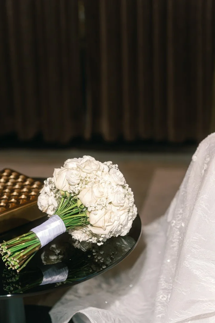 A bouquet of white roses and baby's breath on a reflective black surface with bridesmaid dress fabric nearby.