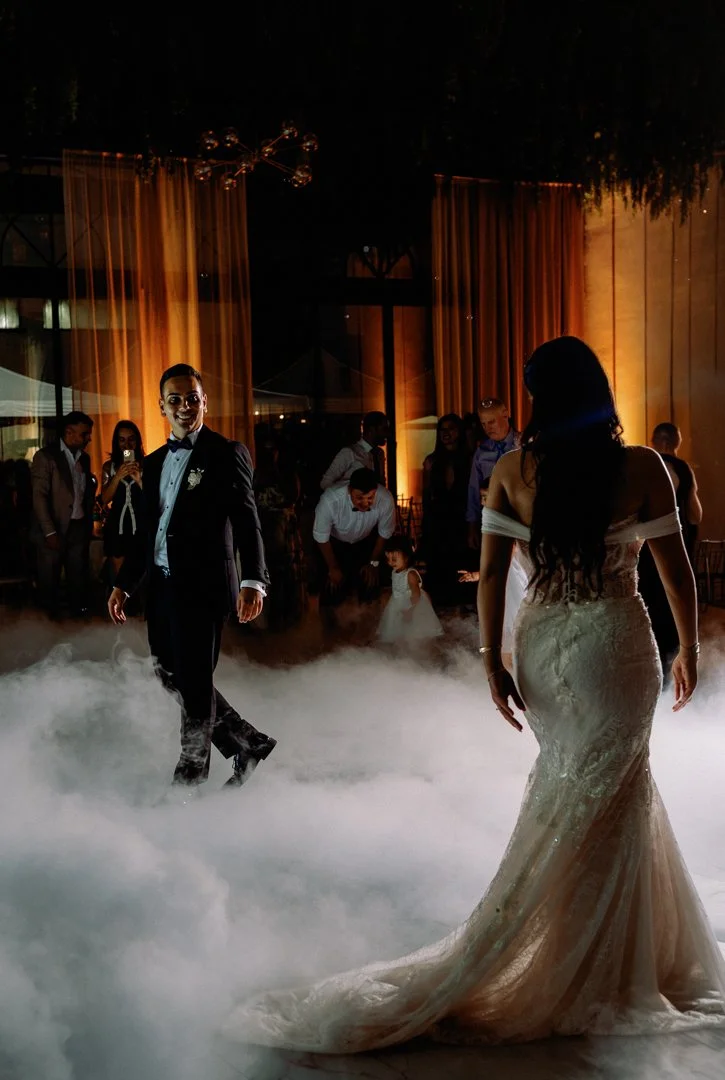 Bride and groom dance at their wedding reception with a foggy dance floor and guests watching in the background.