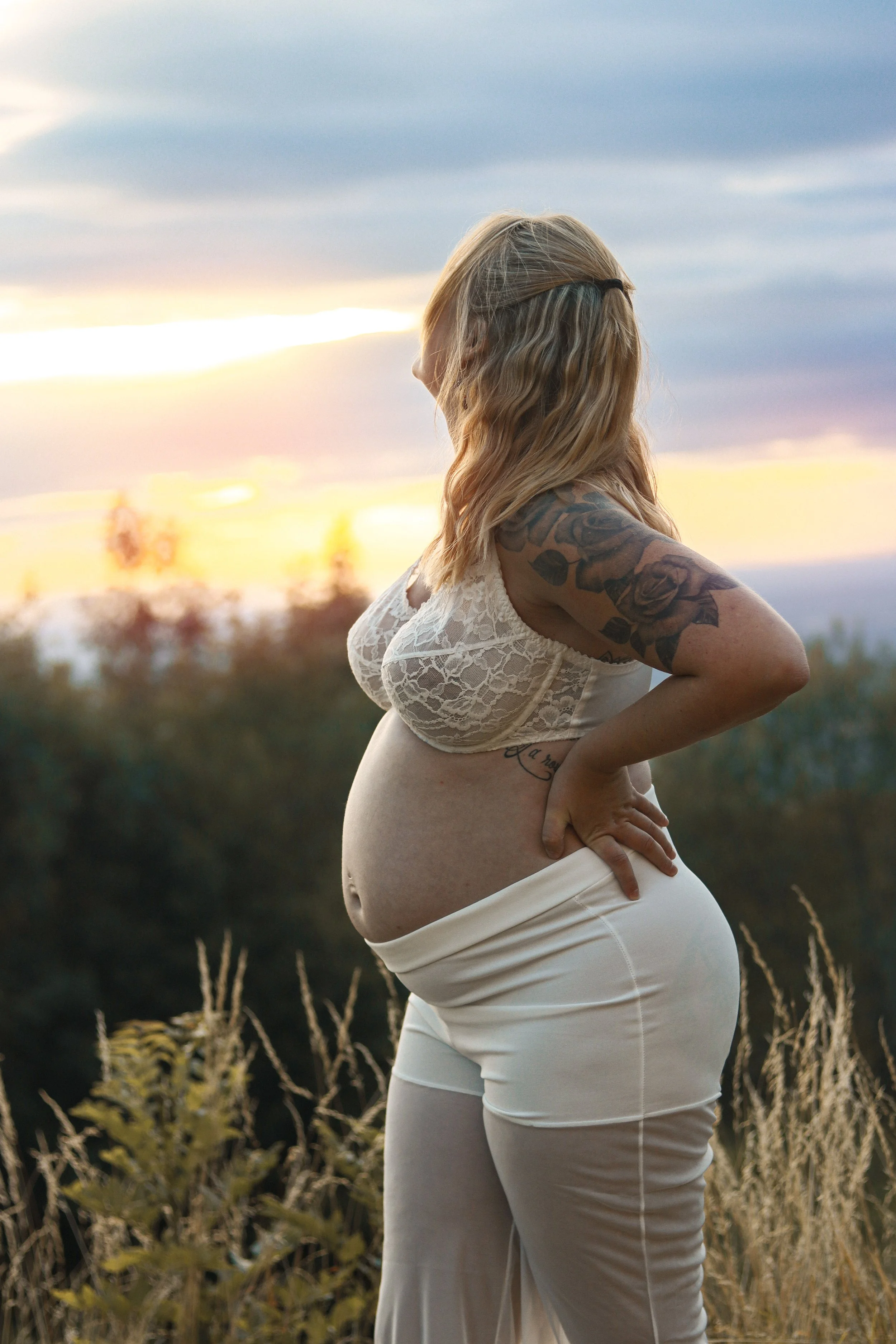 Une femme enceinte en position de profil dans une nature avec un coucher de soleil à l'arrière-plan, portant une robe en dentelle claire et ayant un tatouage sur le bras.