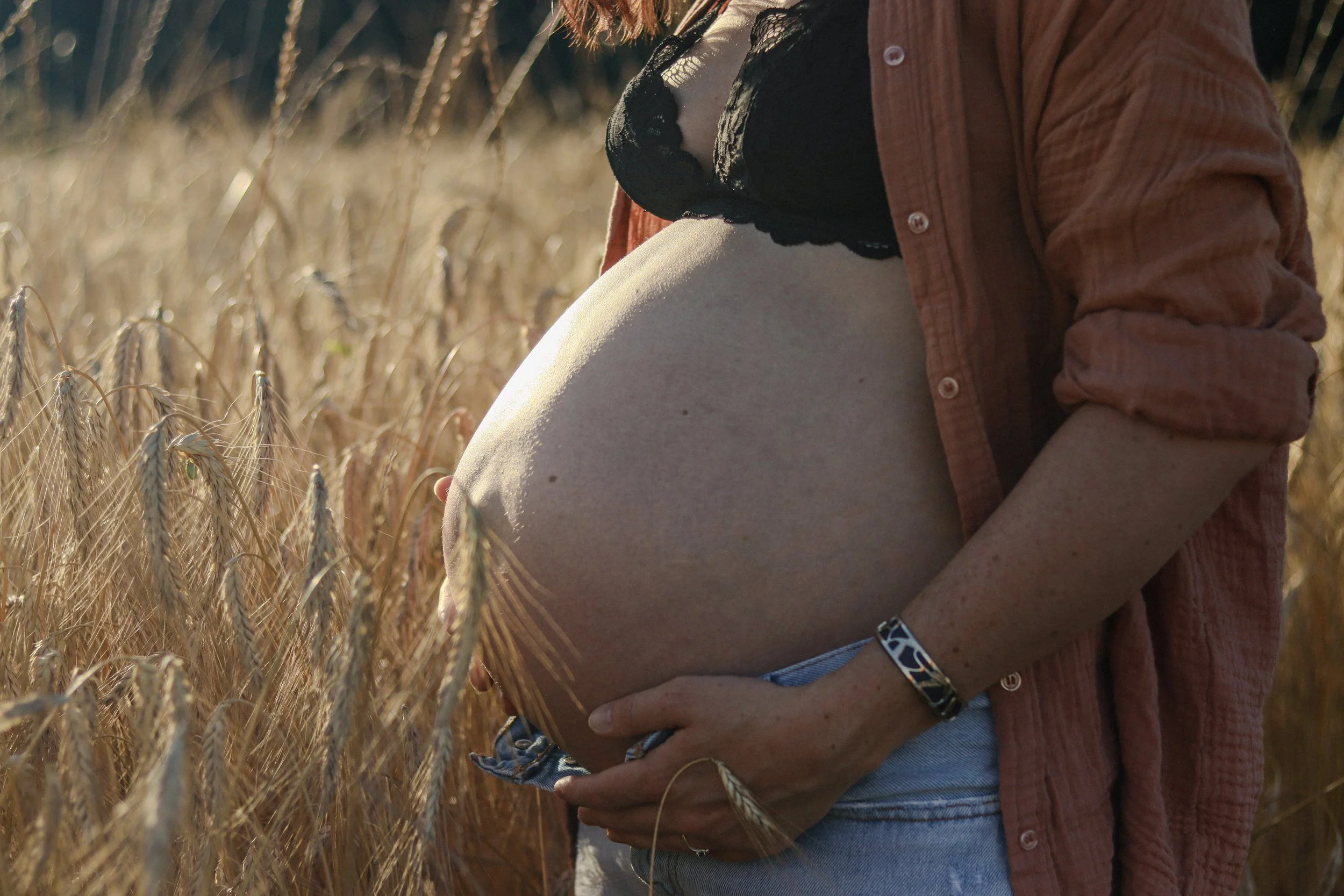 Une femme enceinte portant un haut noir en dentelle, une chemise en flanelle beige, et un bracelet, se tient dans un champ de blé