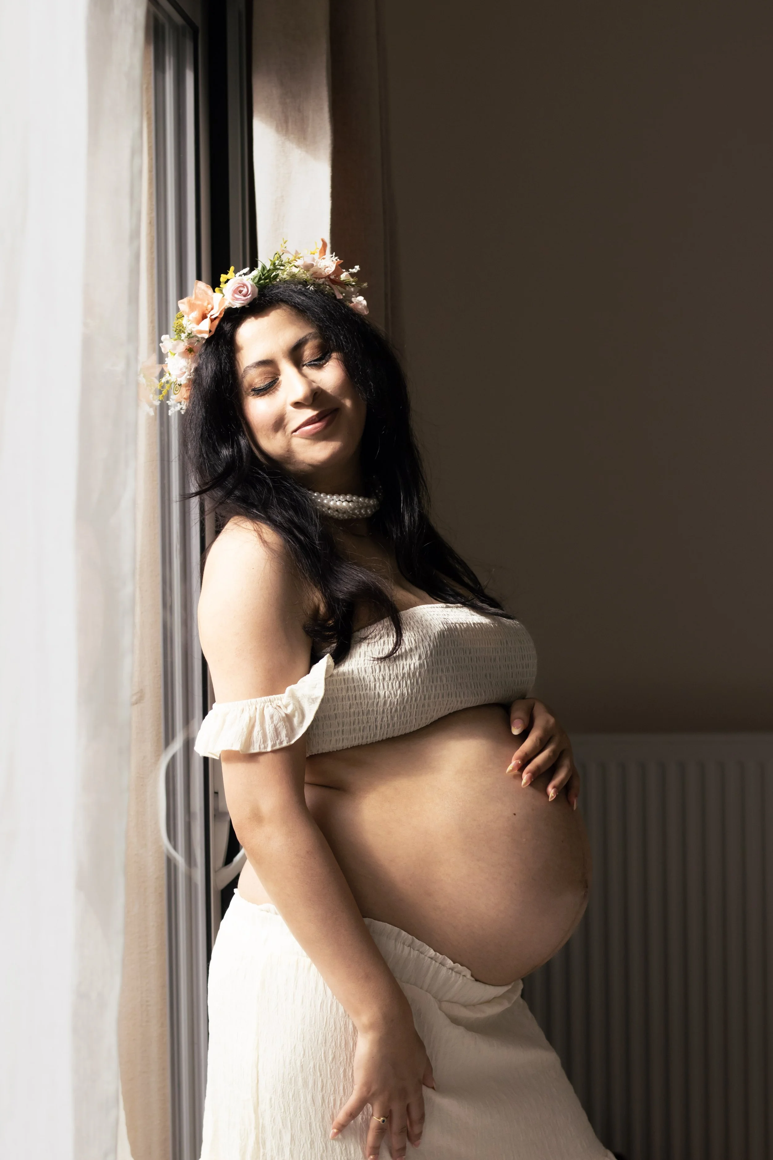 Femme enceinte portant une robe crème, avec un collier de perles, portant une couronne de fleurs pastel, se tenant près d'une fenêtre avec le soleil qui illumine son visage, dans une pièce intérieure.