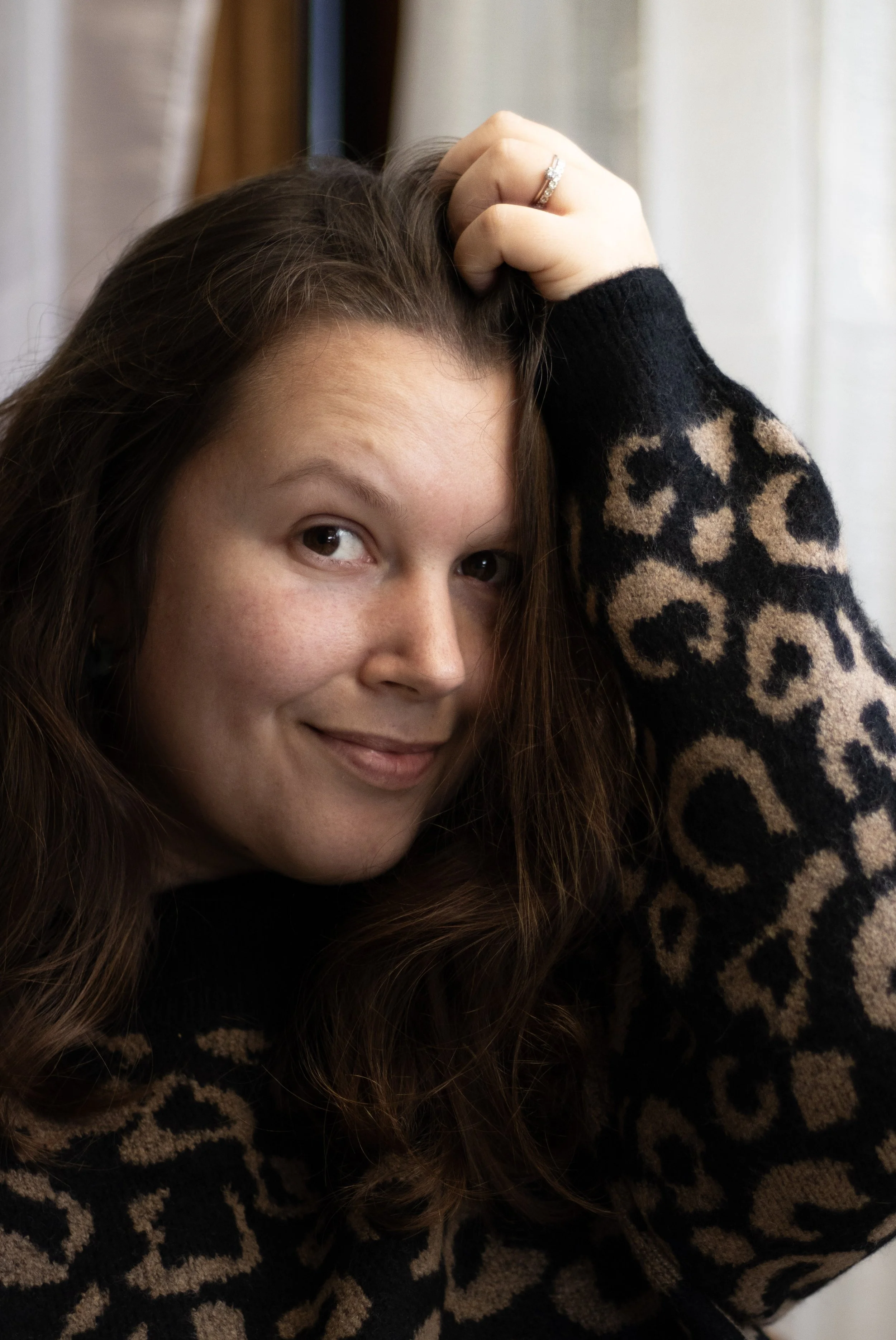 Jeune femme souriante avec les cheveux longs, portant un pull à motif léopard, posant près d'une fenêtre.