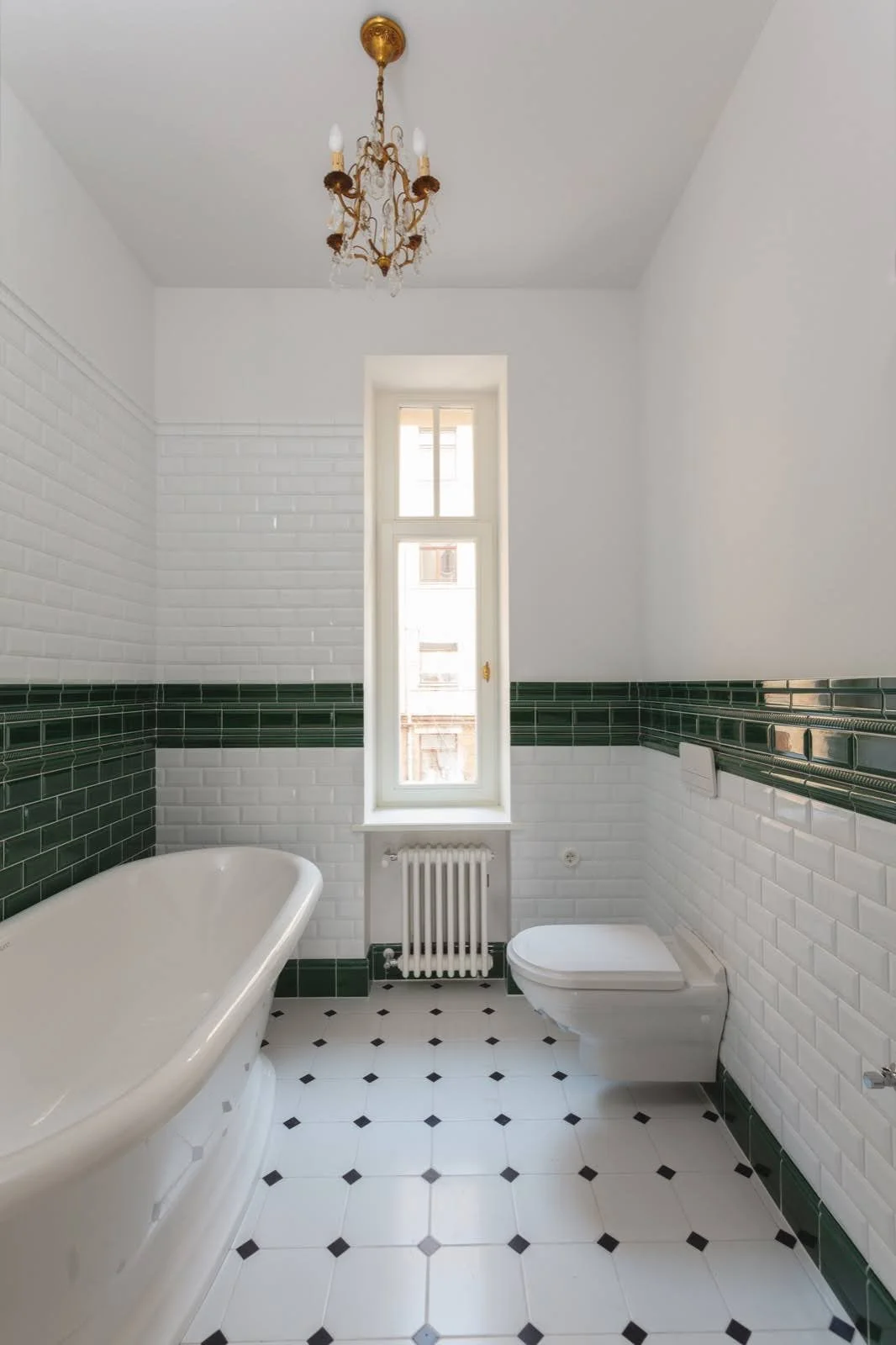 Bathroom with white tiled walls, green tile accent border, black and white checkered floor, a bathtub, a toilet, a tall window, a radiator, and an ornate chandelier hanging from the ceiling.