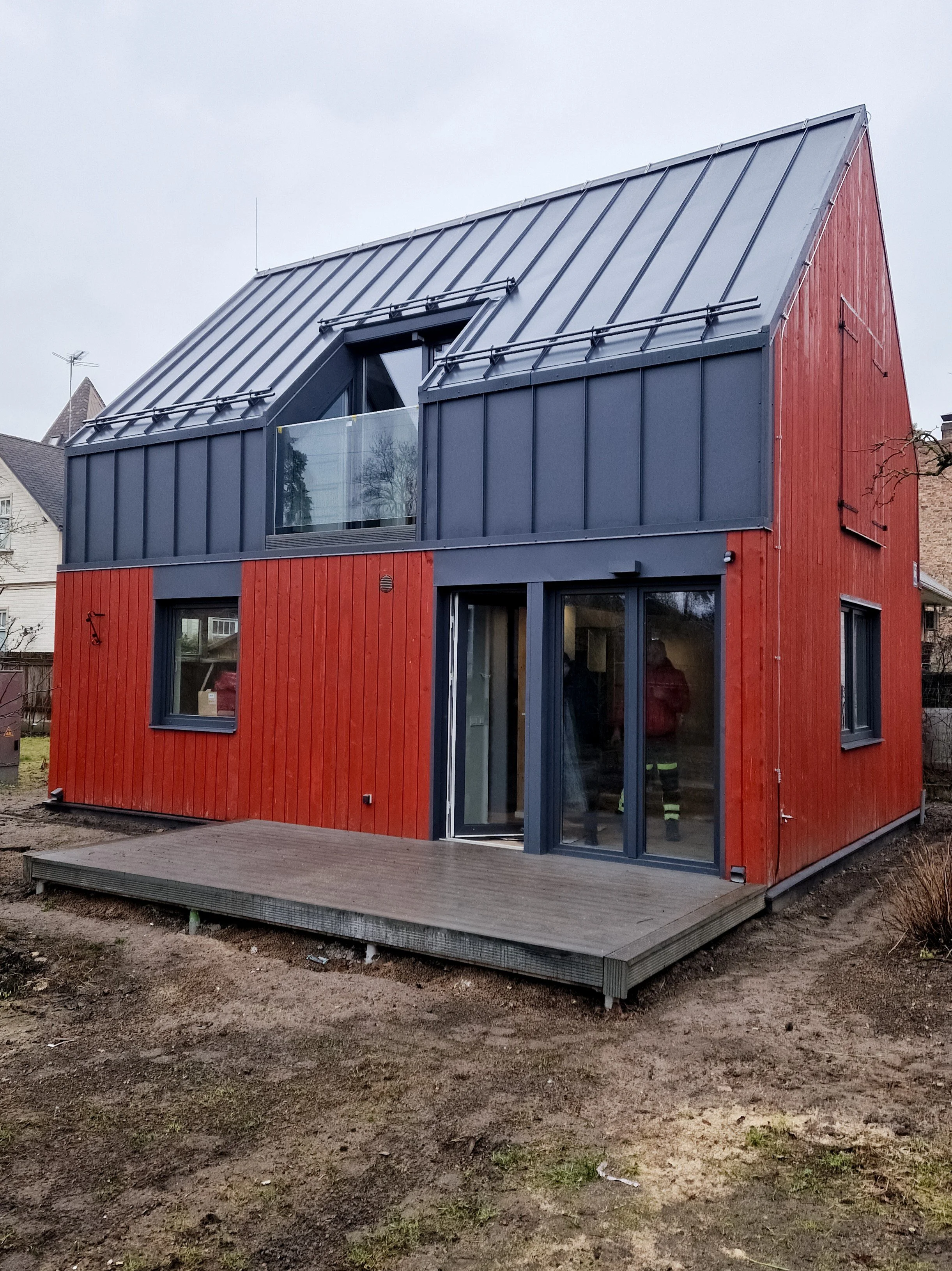A modern two-story house with red wooden siding and a metal roof, featuring a balcony and large sliding glass doors.