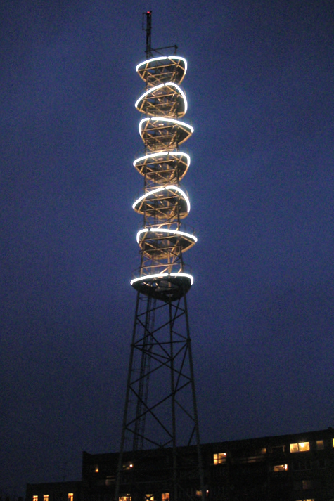 A tall radio transmission tower with spiraling illuminated bands around its upper section against a dark evening sky.