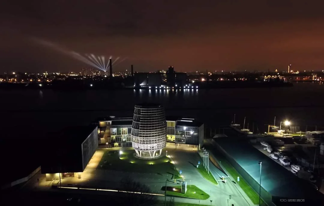 Night view of a modern building with a rounded glass tower, illuminated surrounding area, and a body of water in the background with industrial skyline and smokestacks emitting lights and fumes.