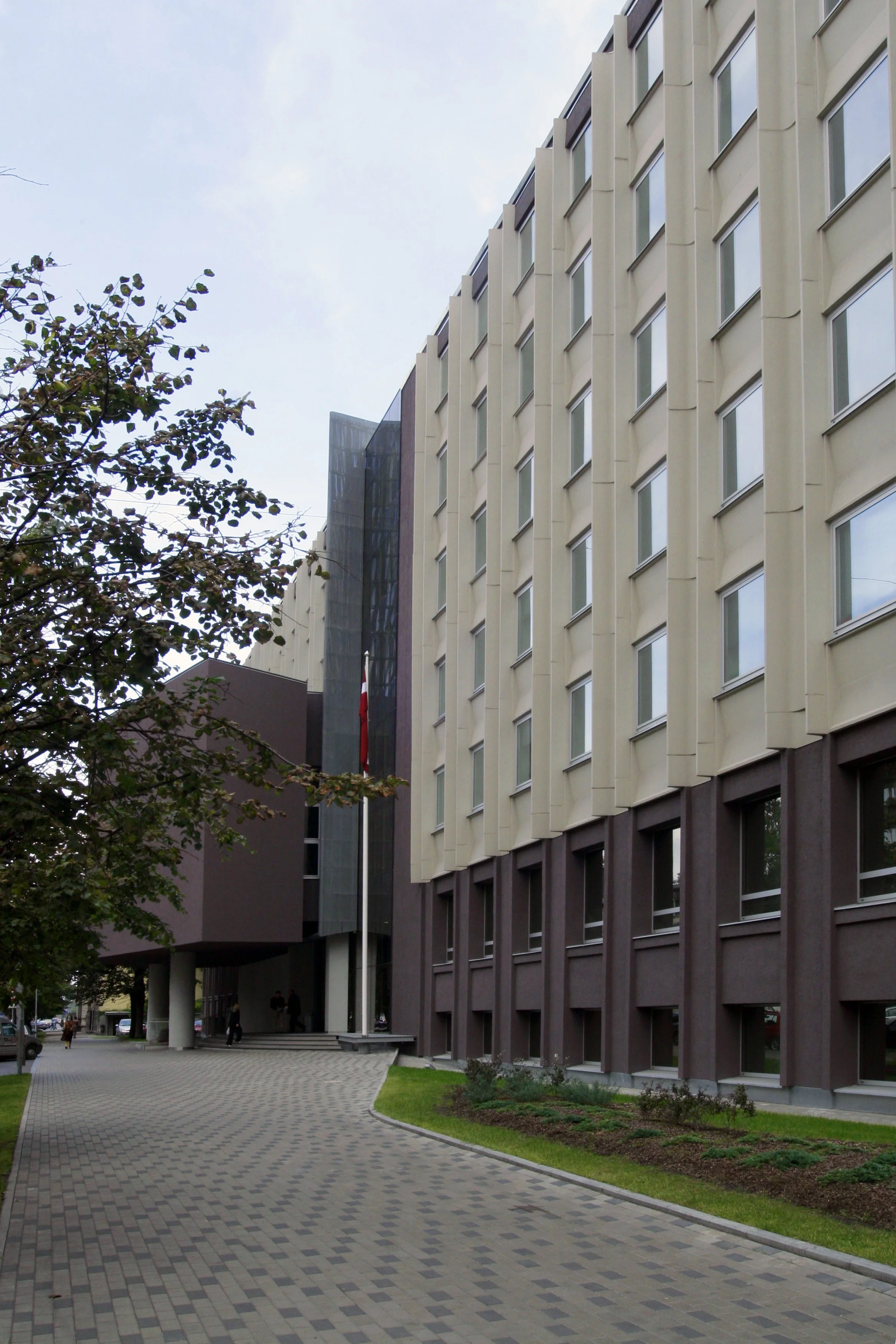 Exterior view of a modern multi-story office building with beige and brown panels and numerous windows, a paved walkway leading up to the entrance, and a tree with green leaves on the left side.