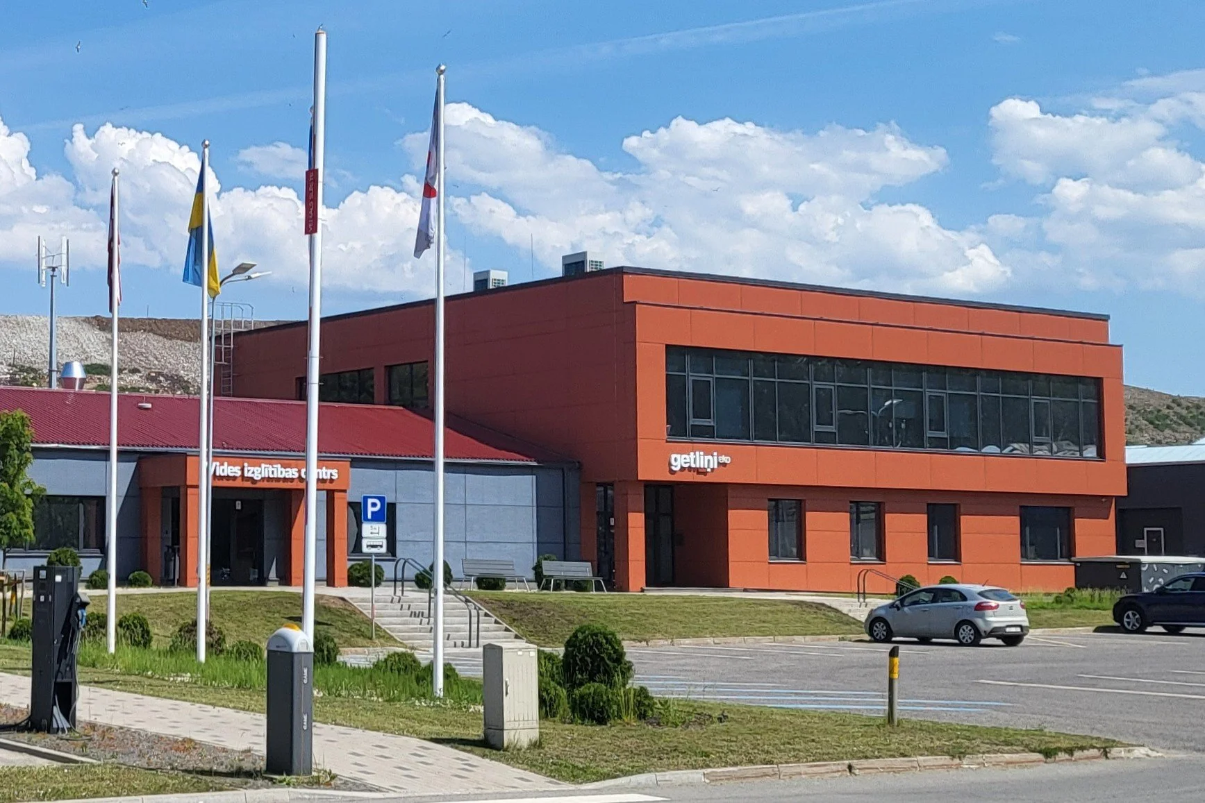 A modern building with a red and gray exterior, labeled 'Vides izglītības centrs' and 'getlini'. There are several flagpoles with flags, a parking lot with cars, and a few trees and bushes in the area.