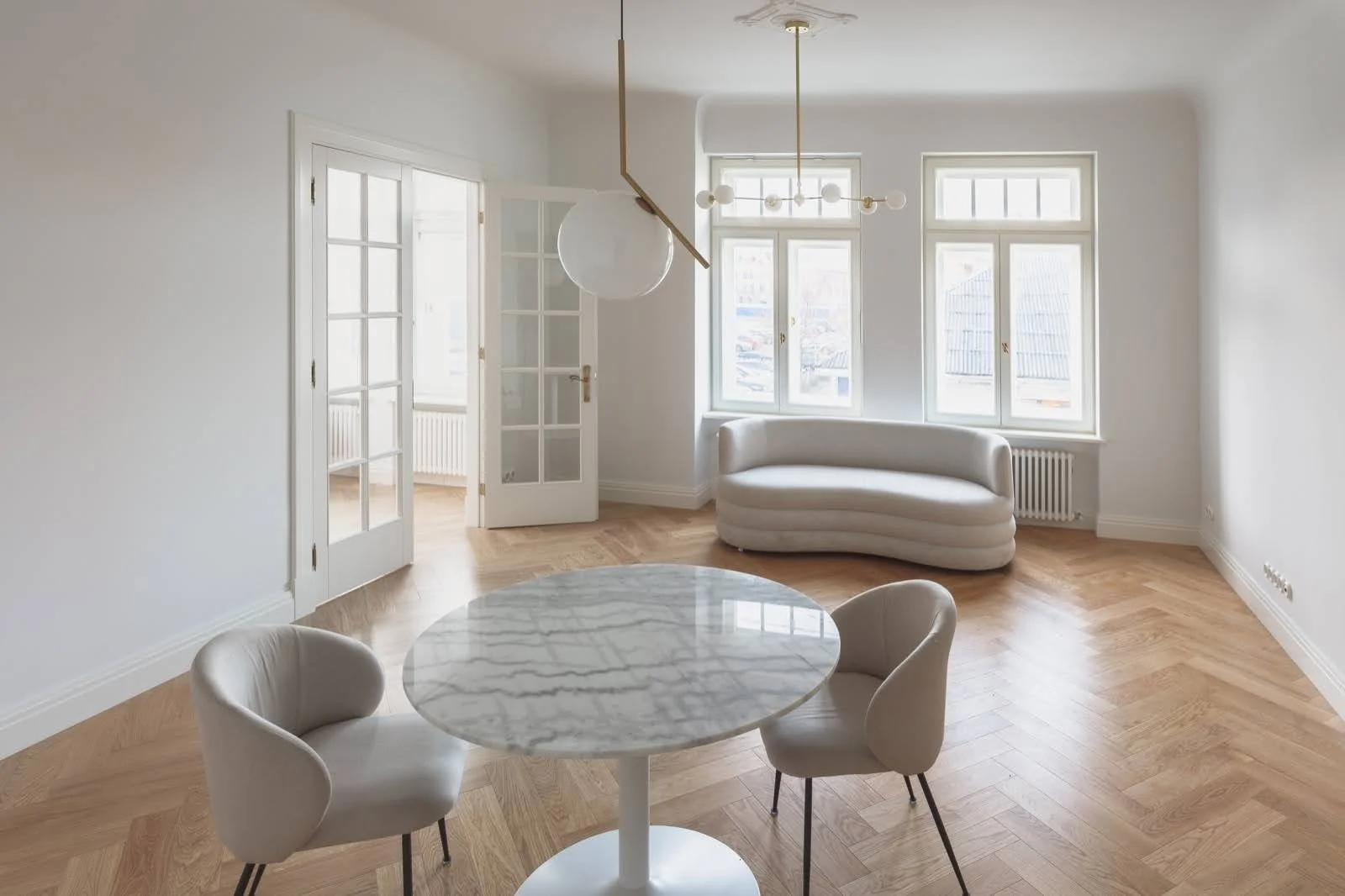 Empty living room with white walls, four large windows, a white curved sofa, a round marble dining table, and two modern beige chairs on a light wooden floor.