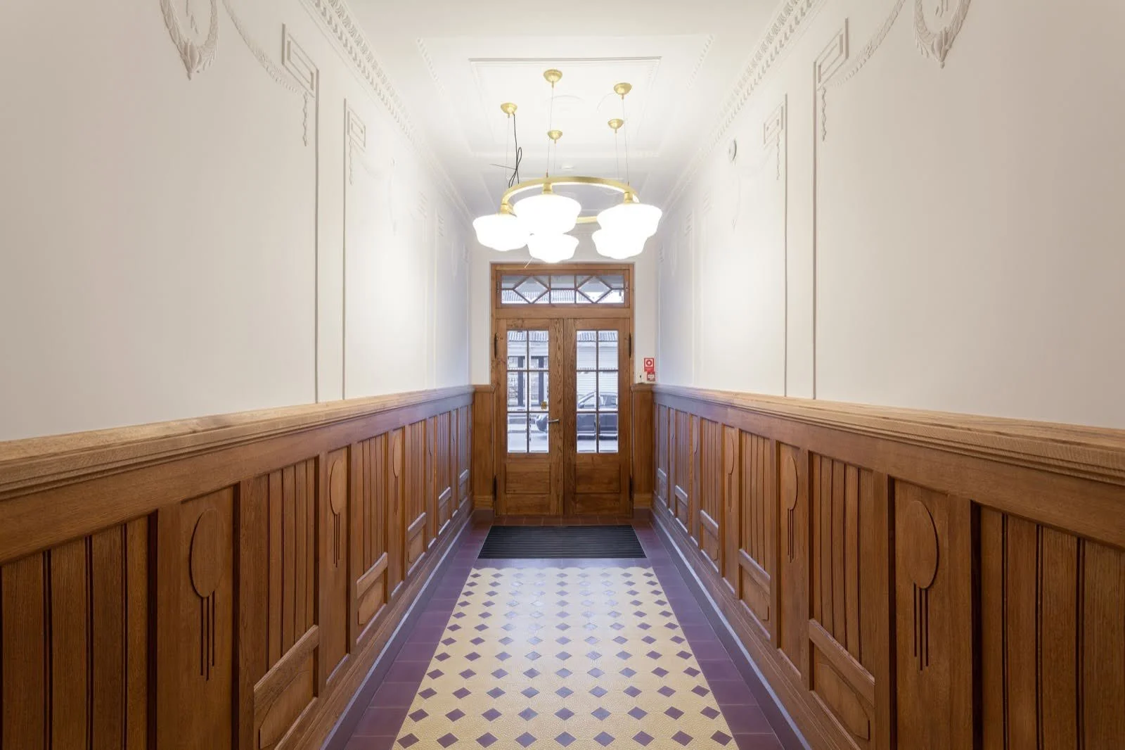 A hallway with white painted walls, wood-paneled wainscoting, a patterned tile floor, and a wooden door at the end, illuminated by a chandelier with multiple white globes.