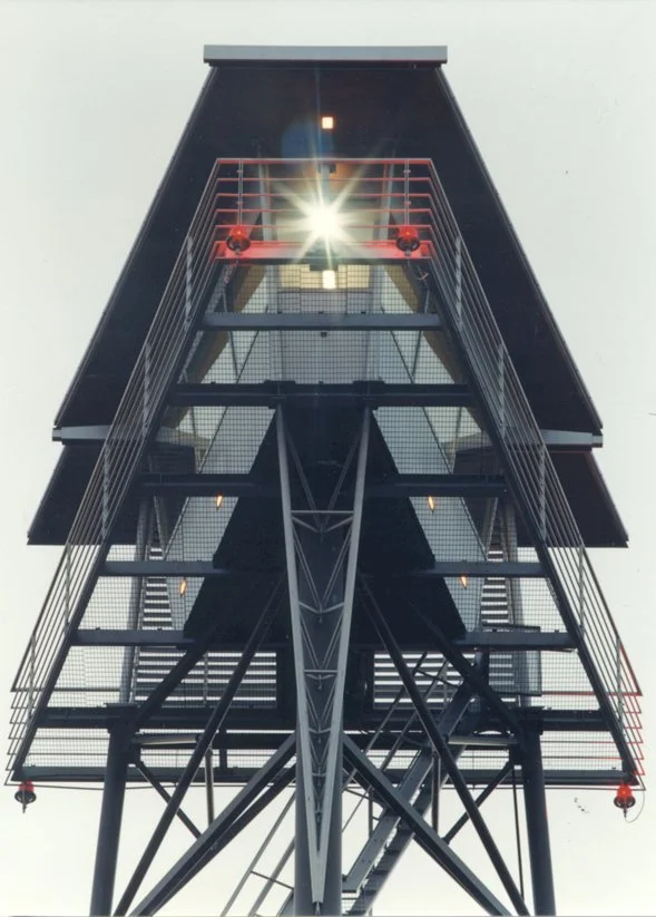 A tall modern fire lookout tower with metal framework and stairs, viewed from below against a cloudy sky.