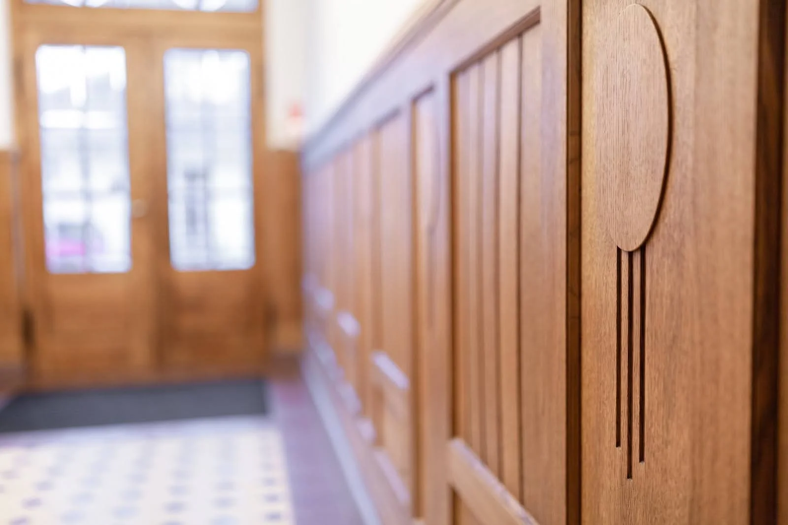 Close-up of stained wood-panel wainscoting with a circular design detail, in a hallway with glass-paneled doors in the background.