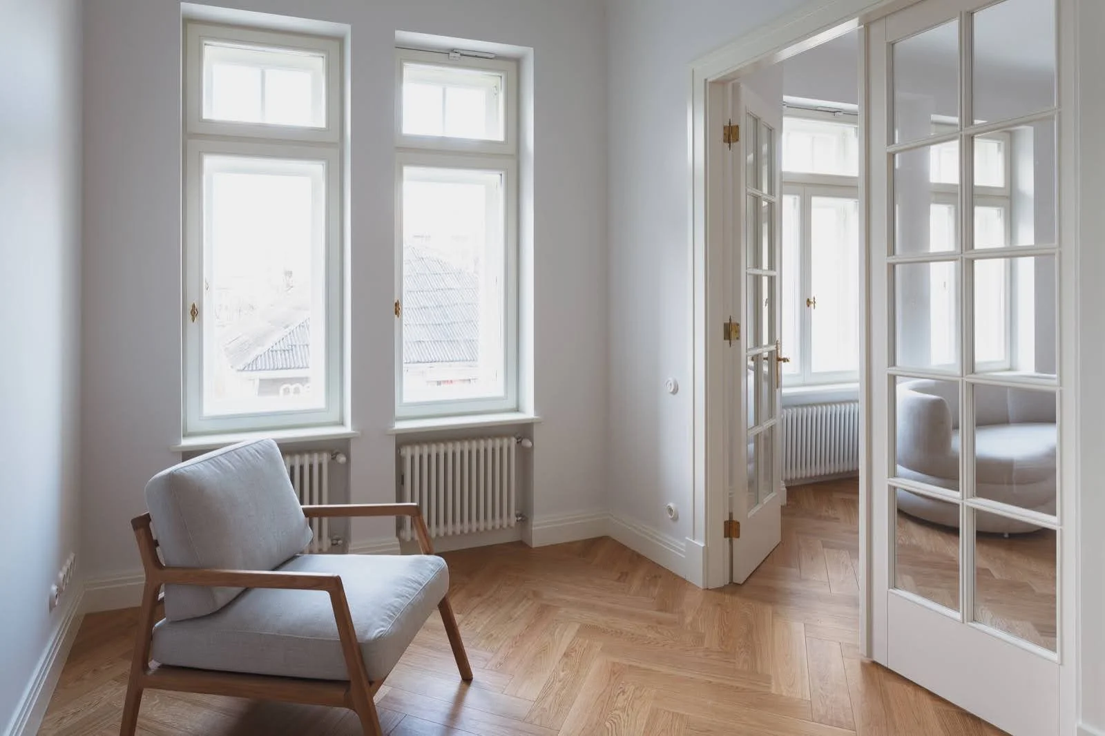 Bright room with large windows, wooden floor, white walls, a cushioned chair with wooden arms, and a glass-paneled door leading to another room.