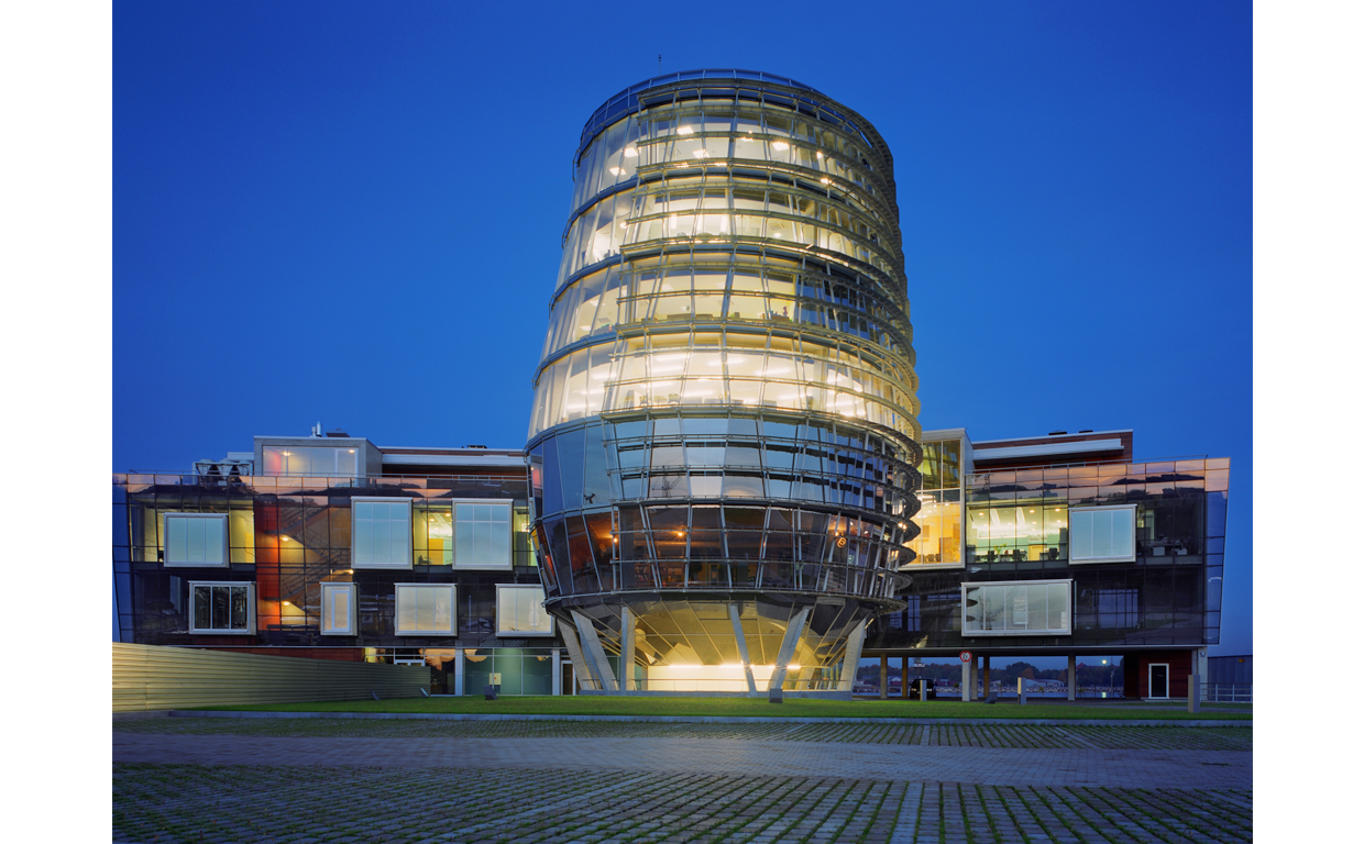Modern multi-story glass building with a rounded tower structure, illuminated from within, during twilight.