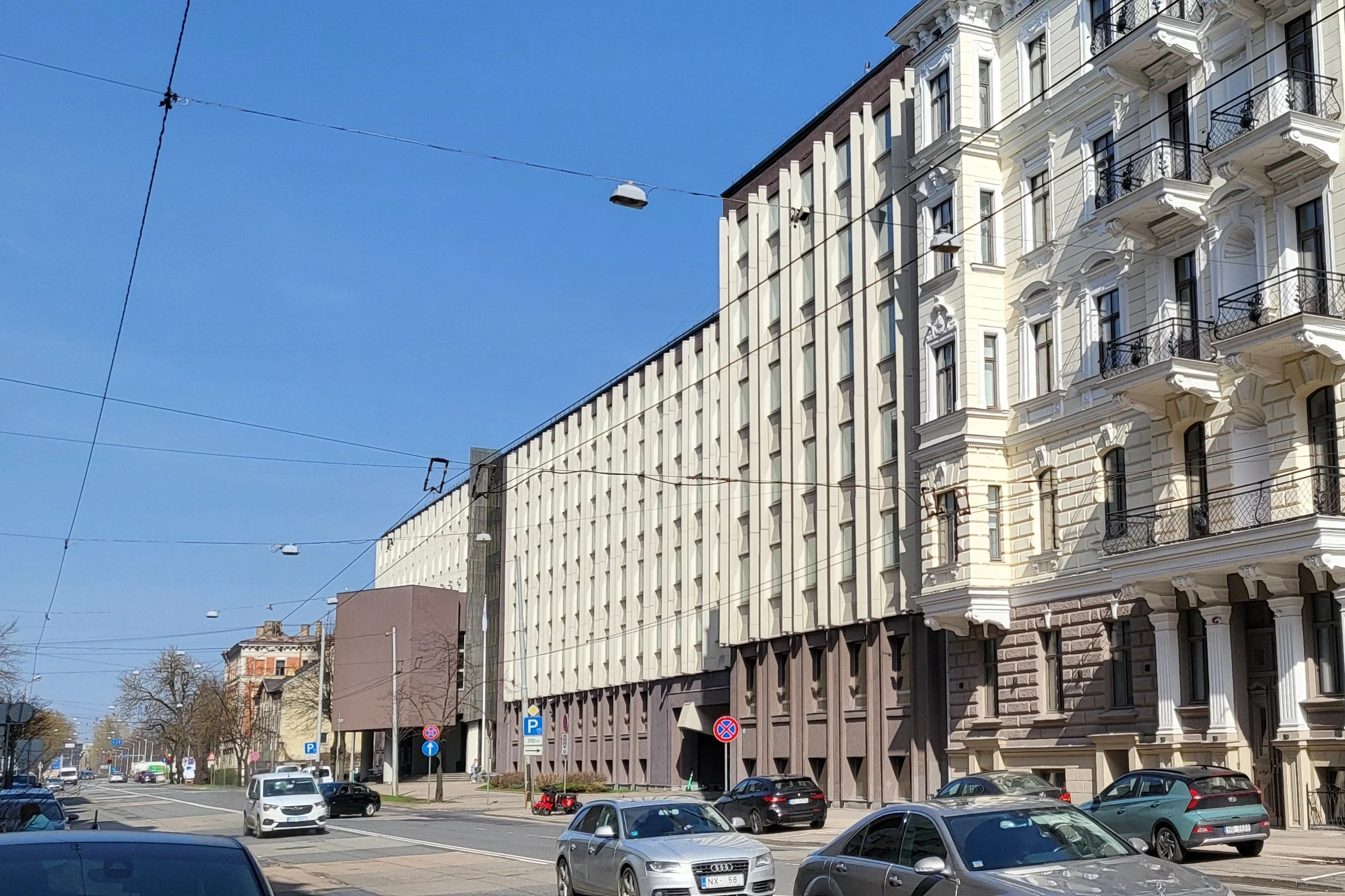 Street view showing parked cars and a mix of modern and historic buildings on a sunny day with overhead tram lines.