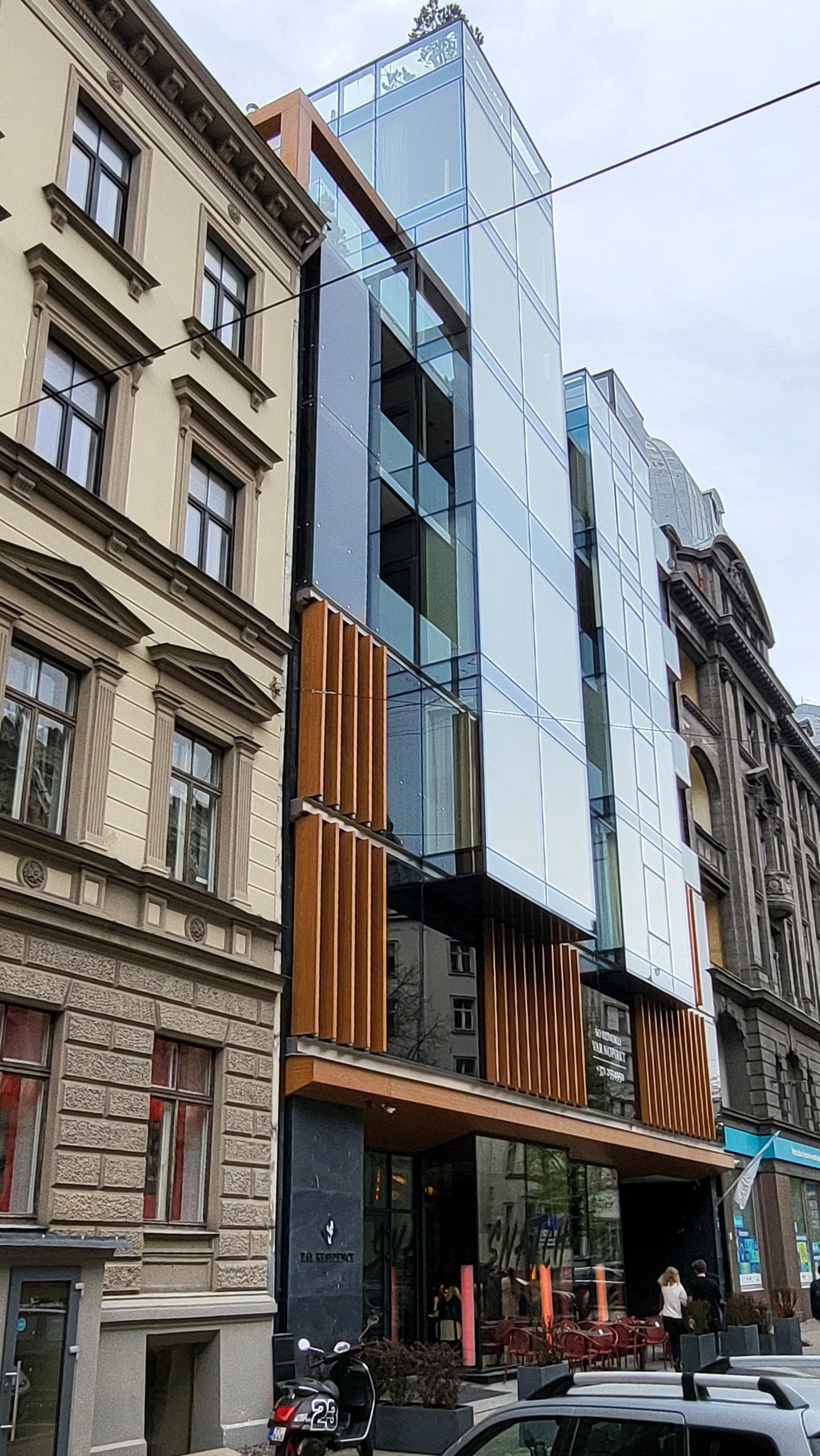 Modern glass building with wooden accents on a city street, adjacent to older stone buildings, with bikes and cars parked outside.