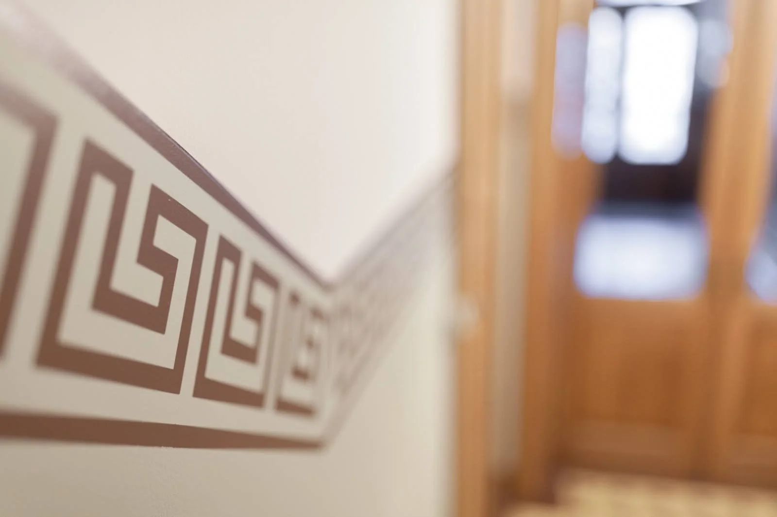 Close-up of wall with decorative trim in a home, showing a geometric pattern in brown and white, with a blurred wooden door and window in the background.