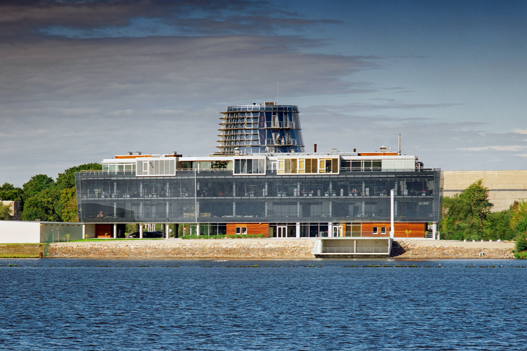 Modern multi-story building with glass exterior on waterfront, surrounded by trees under cloudy sky.