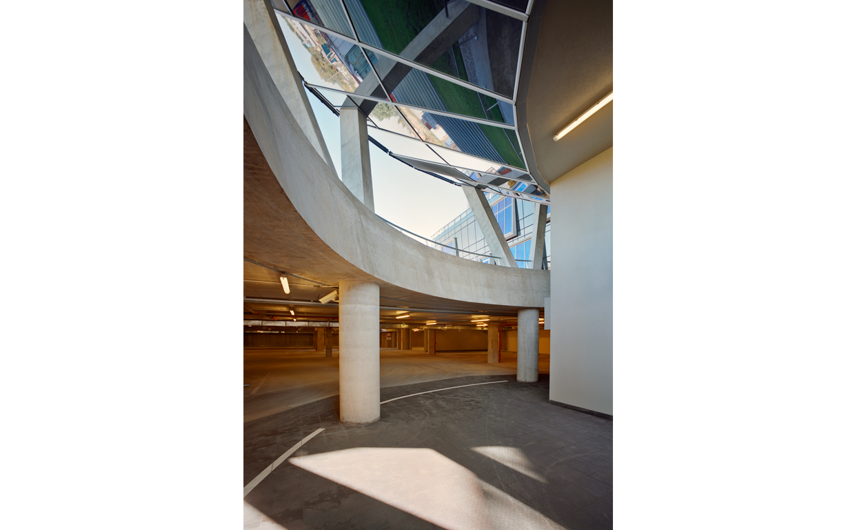 Interior view of a modern parking garage with circular concrete pillars, curved concrete beams, and a large window with reflective glass panels displaying the urban exterior.