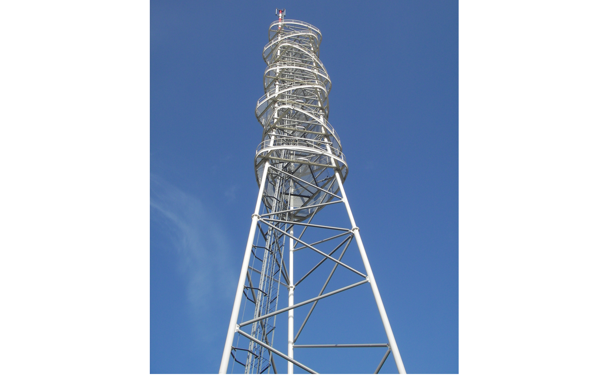 A tall metal communication tower with spiral staircase and safety railing against a clear blue sky.