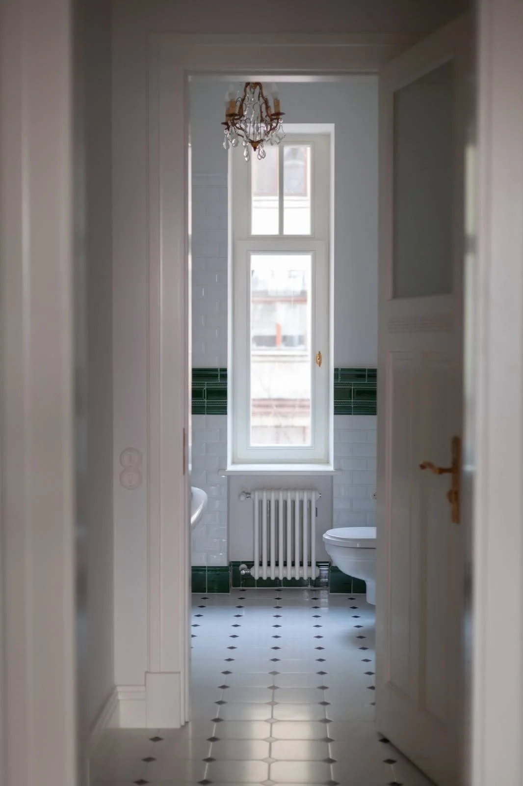 Inside view of a bathroom with a tall window, green and white tiled walls, a radiator, a toilet, and a chandelier hanging from the ceiling.