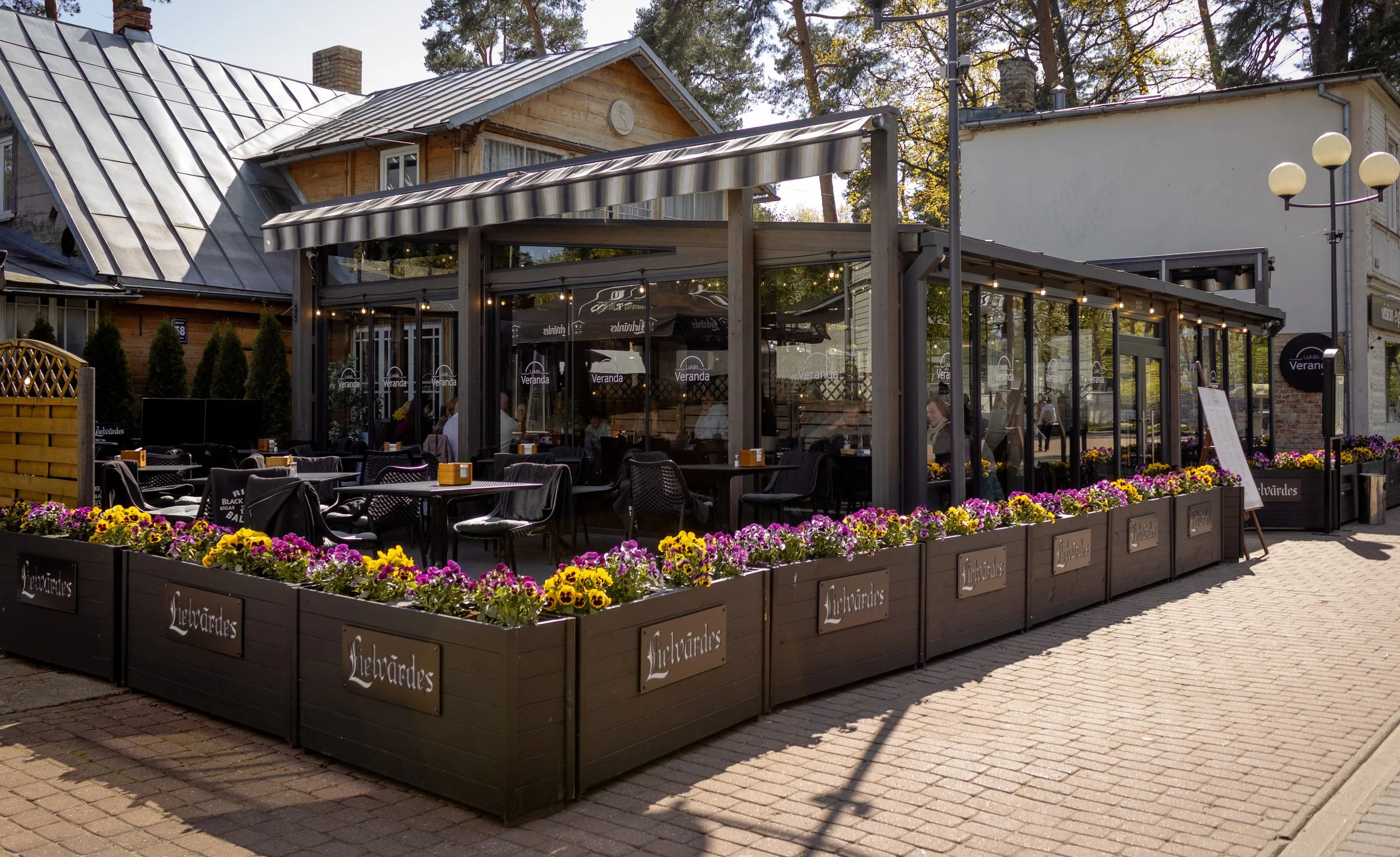 Outdoor restaurant patio enclosed with glass walls, decorated with colorful flowers in dark wooden planters, and furnished with black tables and chairs, with some customers dining inside visible through the glass.