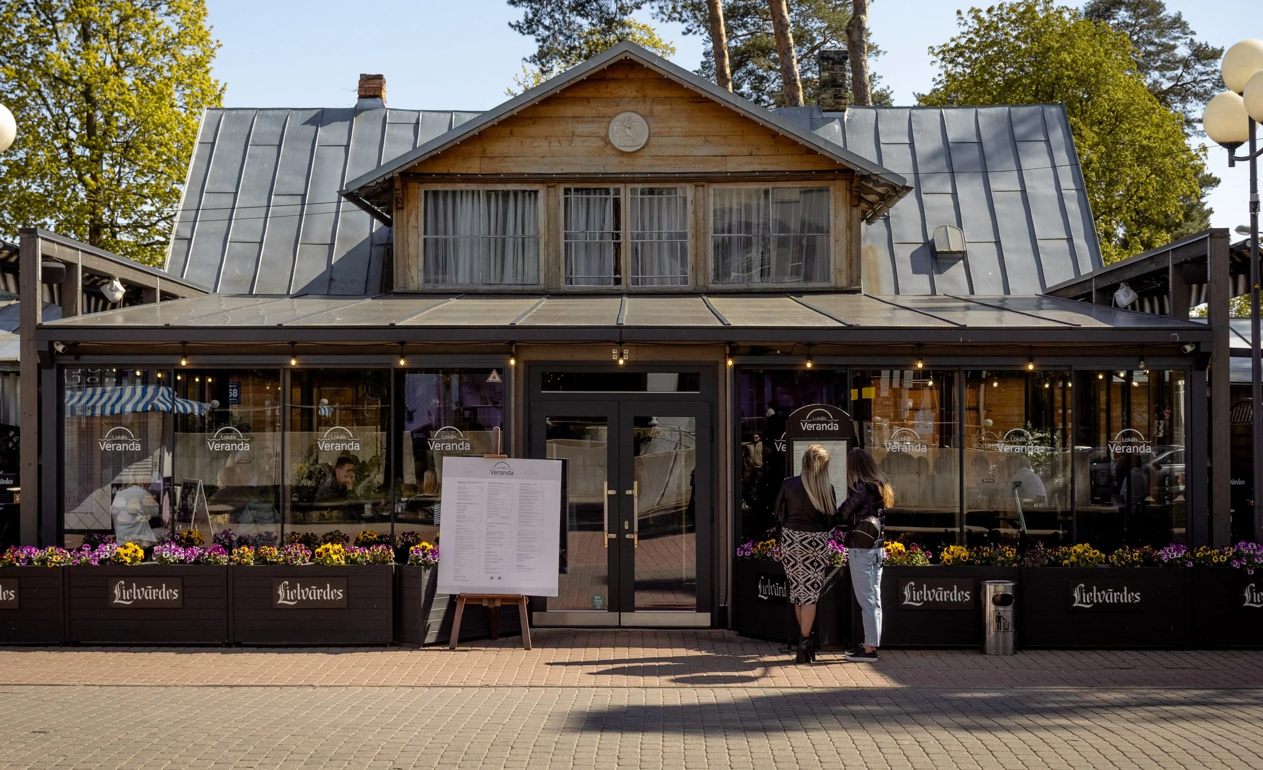 Front view of a restaurant with glass windows, black exterior, and patio seating, with two women standing outside in conversation.
