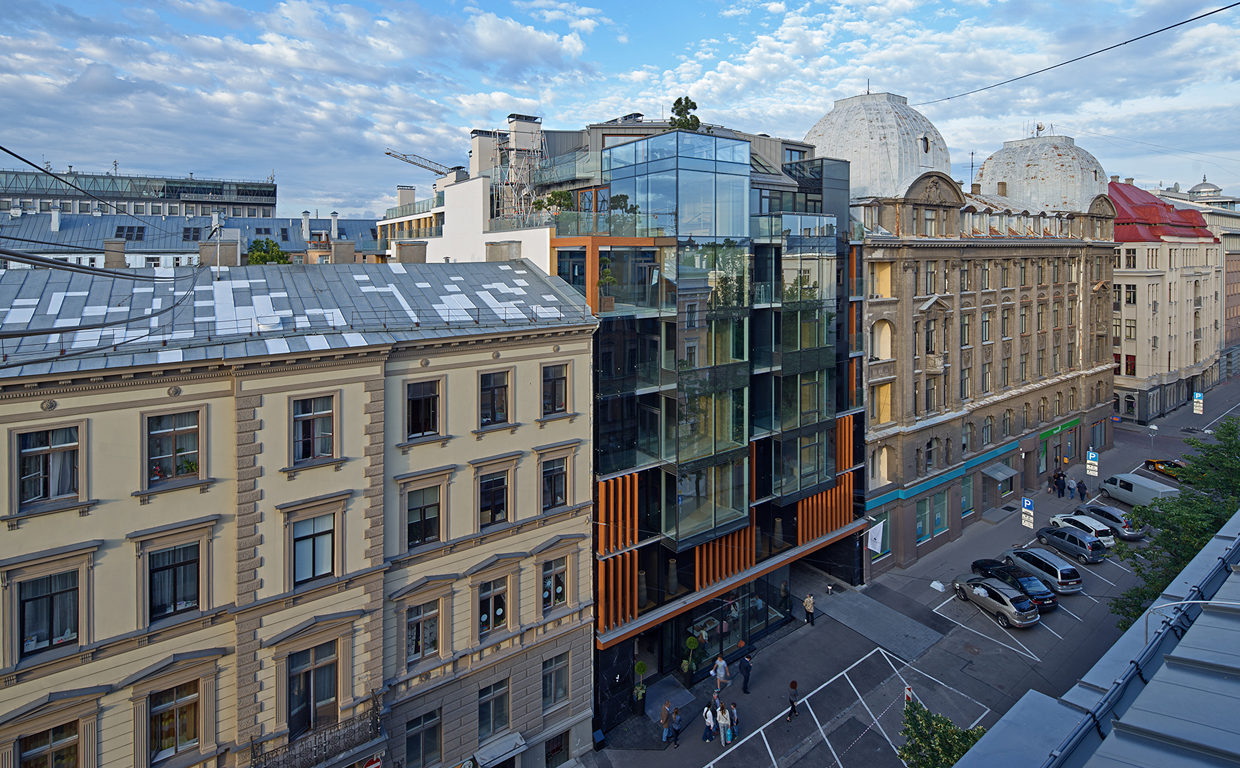A city street with parked cars and pedestrians, modern glass building in contrast to older historic buildings, blue sky with scattered clouds.