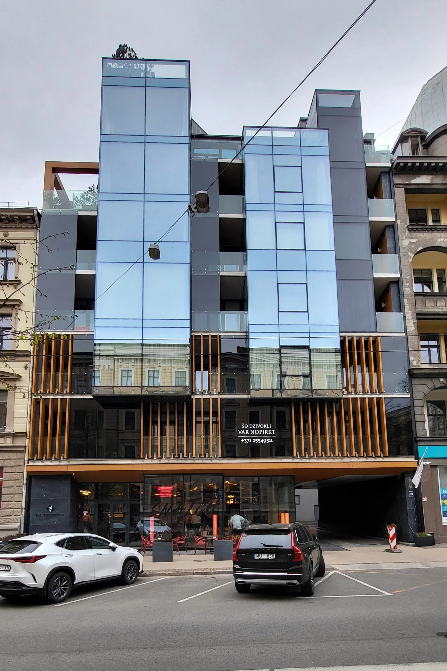 A modern multi-story building with a glass facade and wooden accents, located on a city street with parked cars and a small cafe at the ground level.