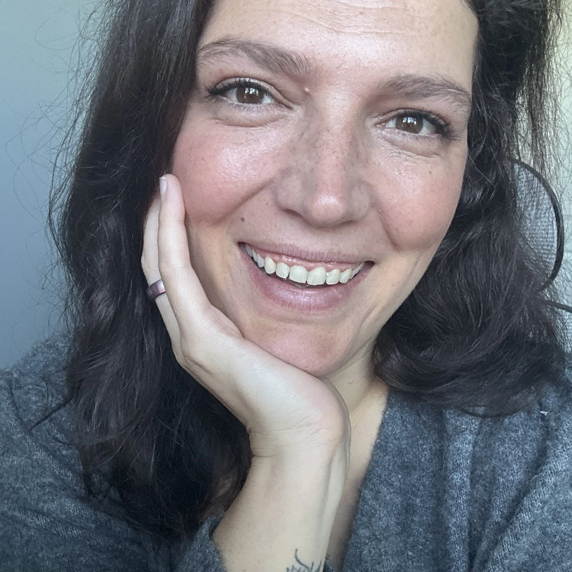 Close-up photo of a woman with curly dark hair, smiling gently, with her hand resting on her cheek, wearing a gray top.