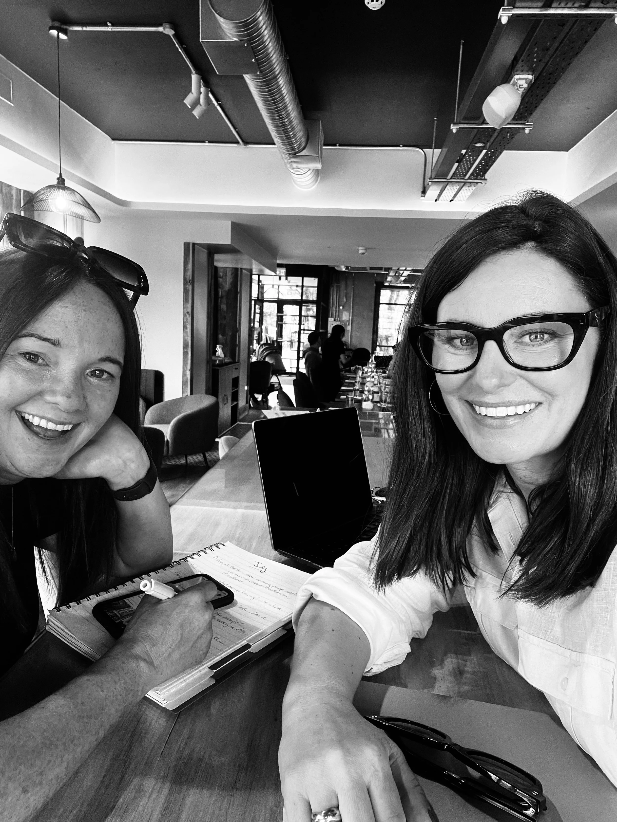 Two women smiling at a table in a modern cafe or restaurant, with notebooks, a laptop, and a smartphone on the table. One woman is wearing glasses and a white shirt, the other has sunglasses on her head. The background shows other people sitting and talking, large windows, and ceiling industrial pipes.