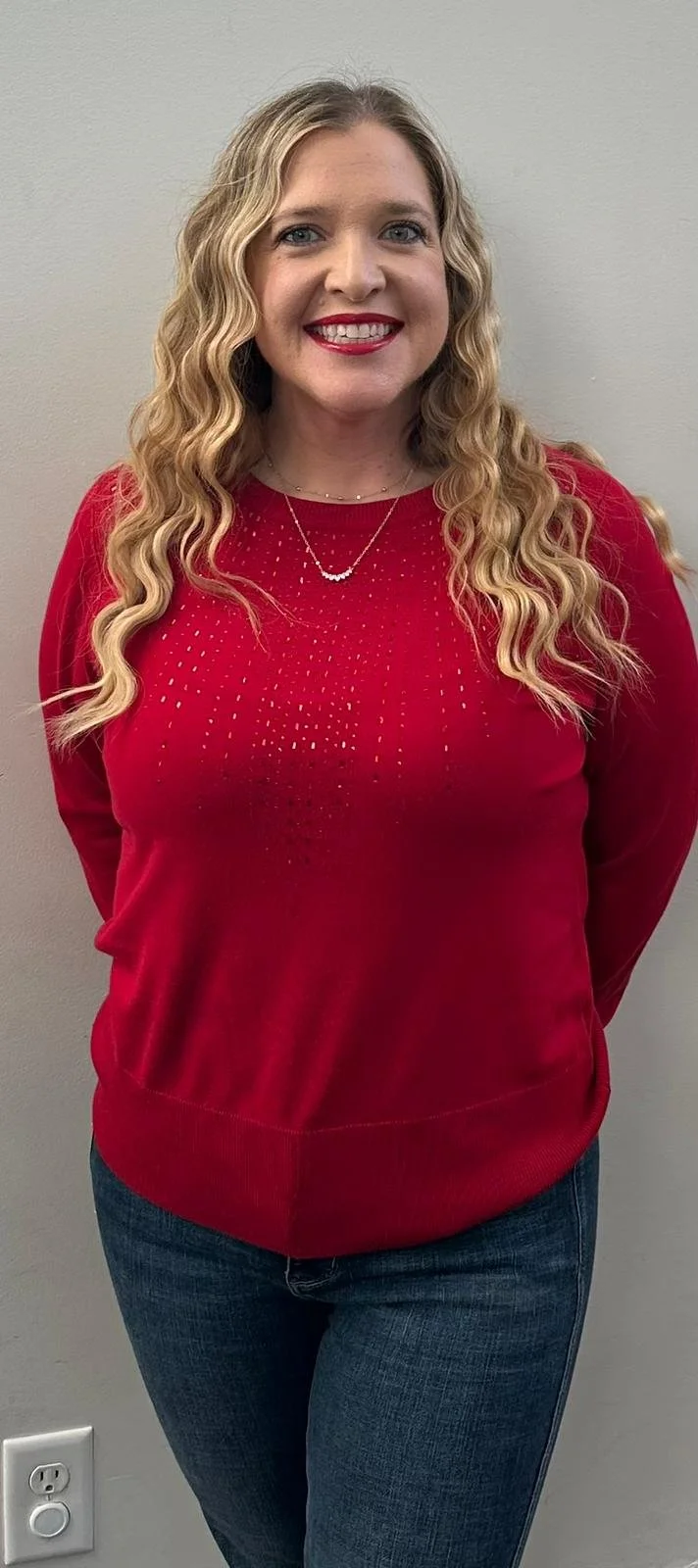 A woman with long, curly blonde hair, wearing a red sweater with small white dots, blue jeans, and a delicate necklace, smiling with red lipstick, standing against a plain gray wall.