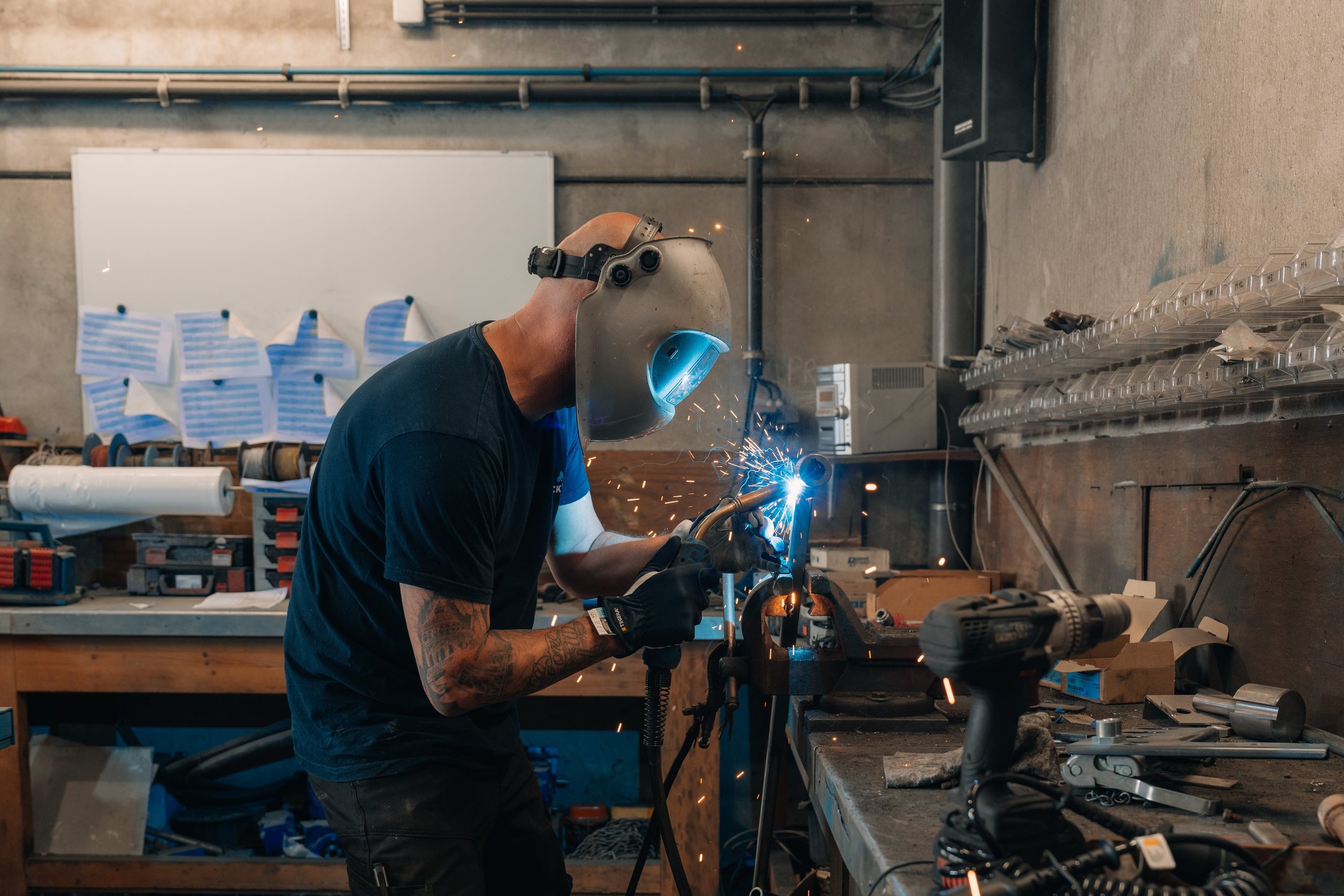 Een man die in een werkplaats een metalen voorwerp aan het lassen is, met veiligheidsuitrusting en gereedschap op de werkbank.