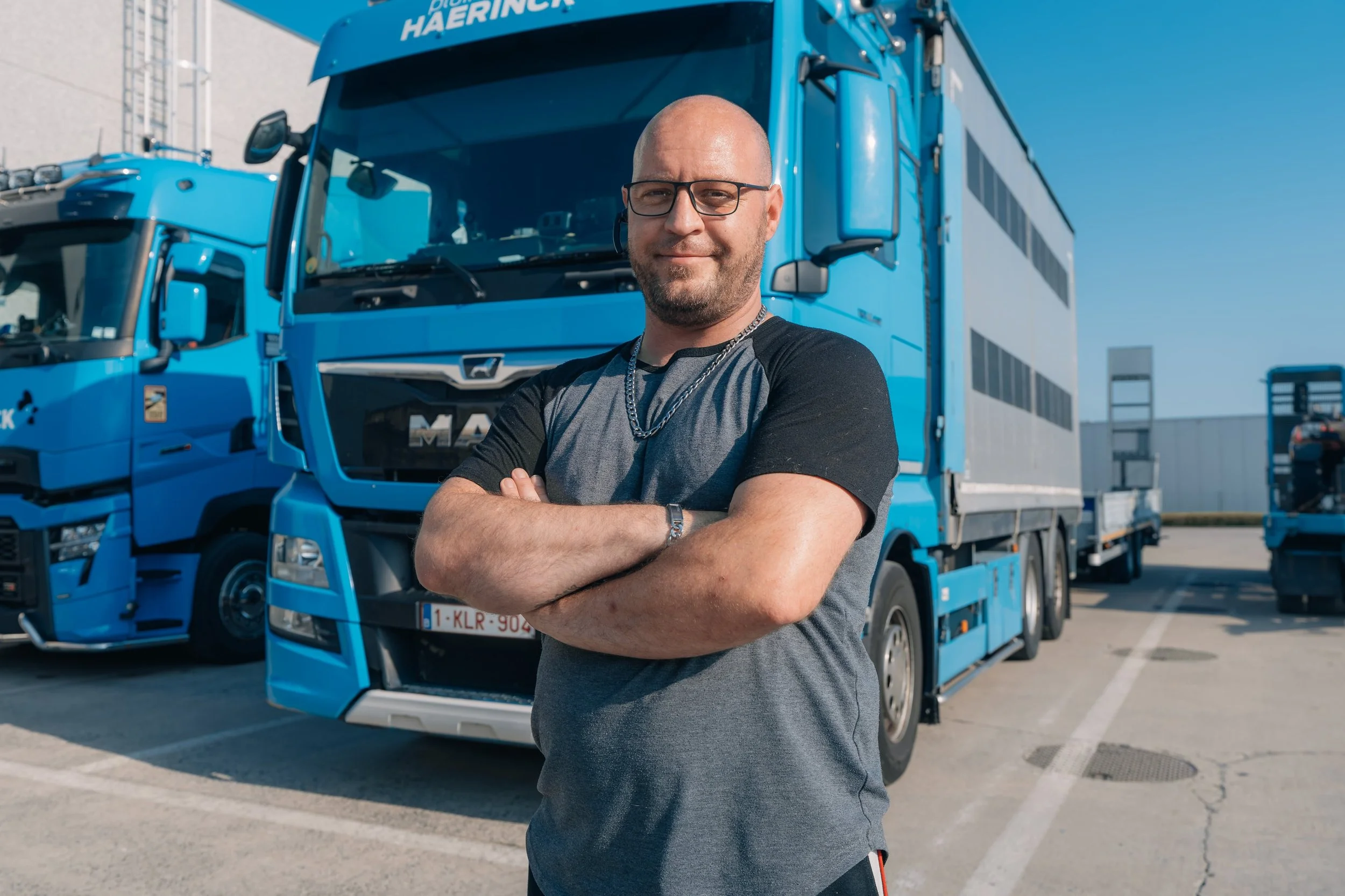 Man met gekruiste armen voor blauwe vrachtwagen op parkeerplaats.