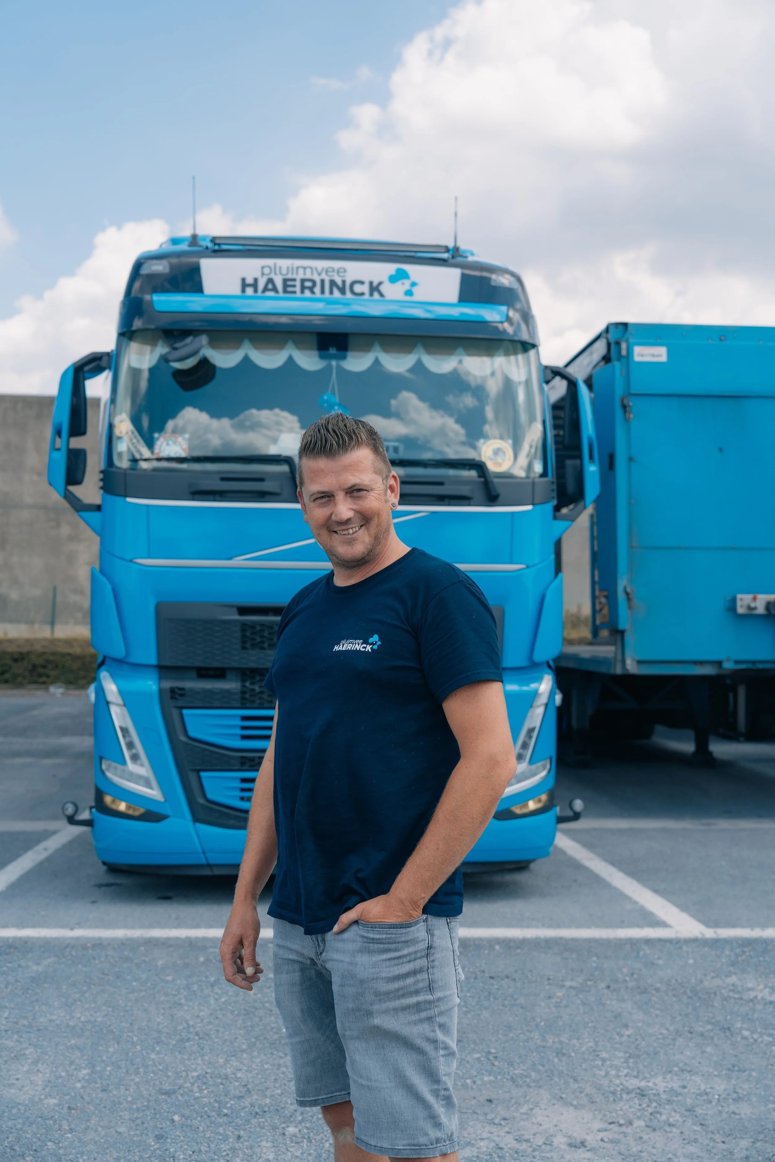 Man staat voor een blauwe vrachtwagen, lachend met hand in zak, op een parkeerplaats.
