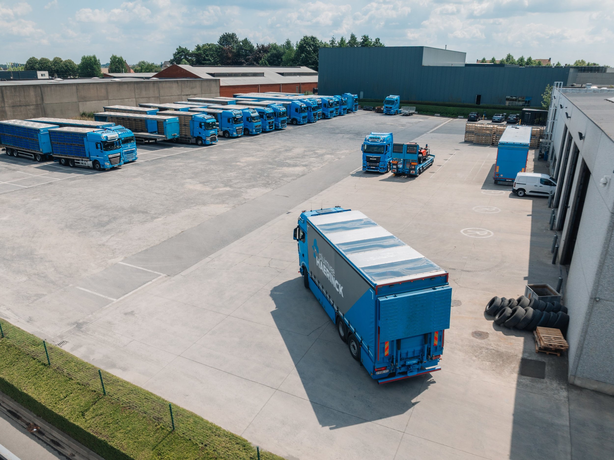 Een opslagplaats met meerdere blauwe trucks en een grote vrachtwagen, enkele kleinere voertuigen, en gestapelde pallets met banden.