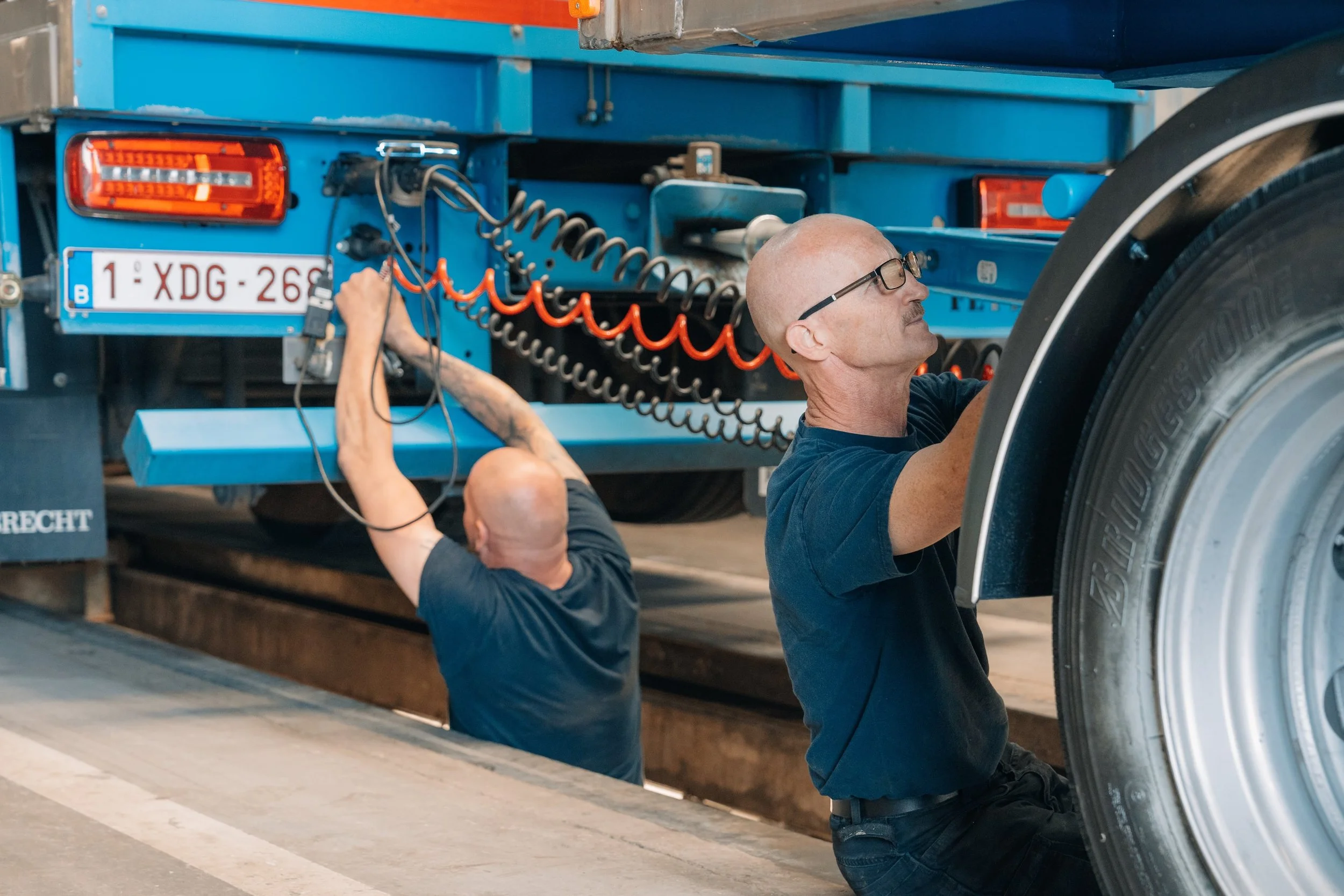 Twee monteurs werken op een grote blauwe vrachtwagen, een zittend en een staand, met gereedschap en elektrische kabels.