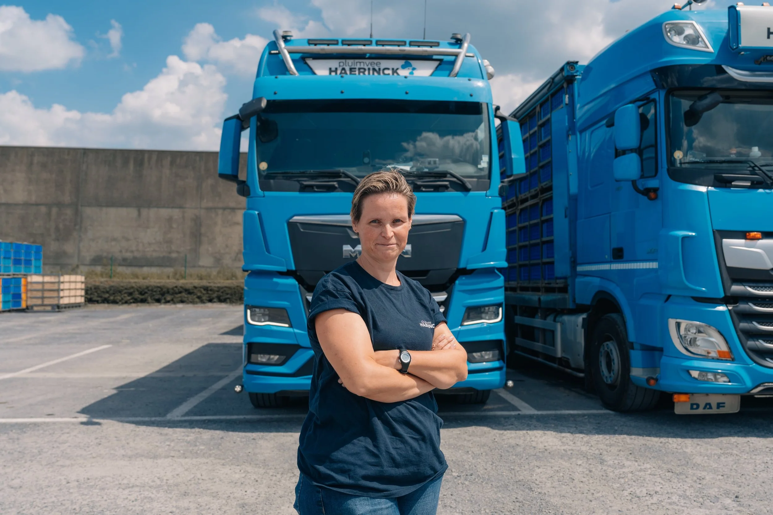 Vrouw staat met gekruiste armen voor twee blauwe trucks op een parkeerplaats onder een bewolkte blauwe hemel.