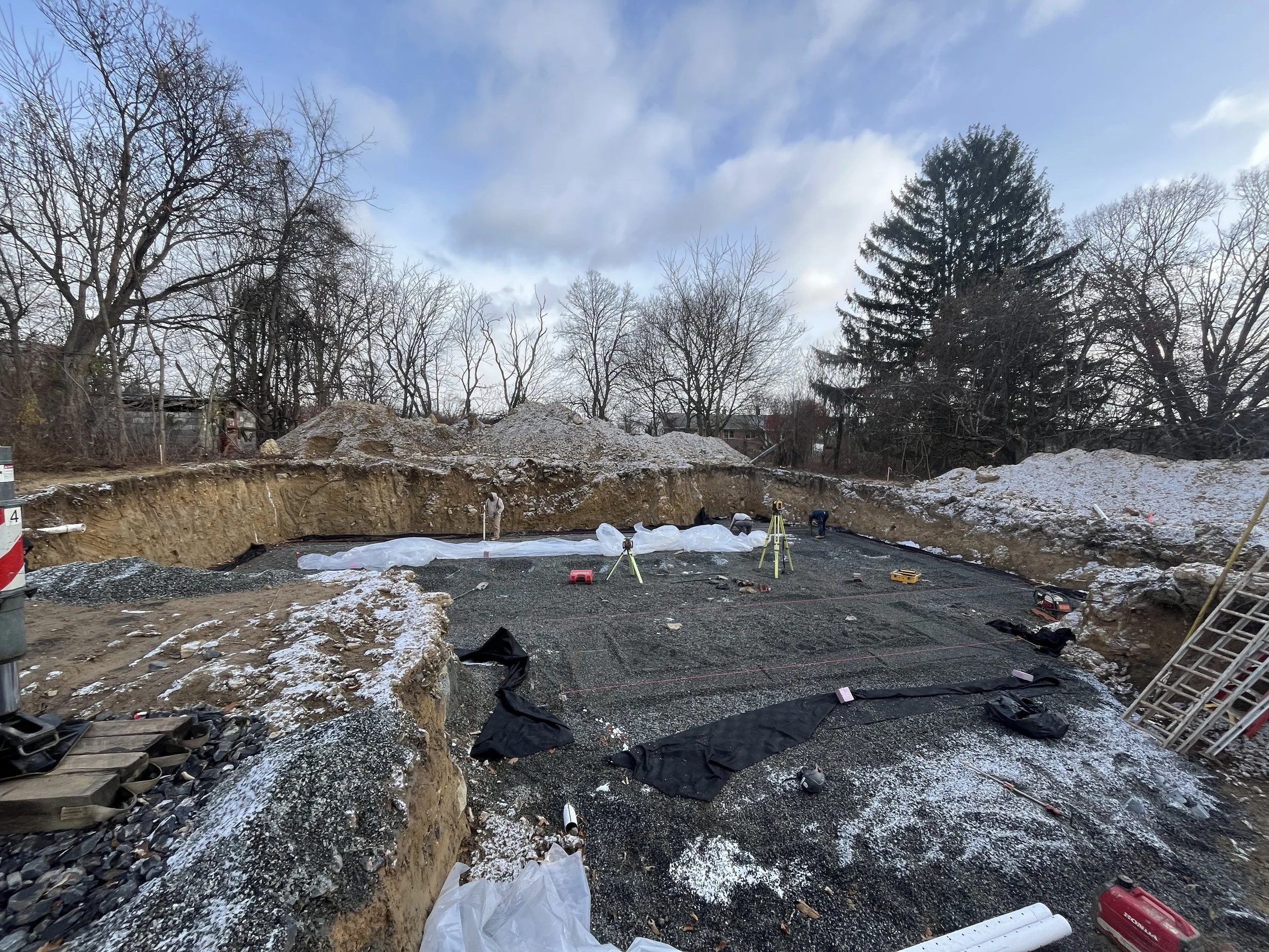 Construction site with a large excavation pit, construction workers, and surveying equipment, late afternoon with partly cloudy sky and leafless trees in background.