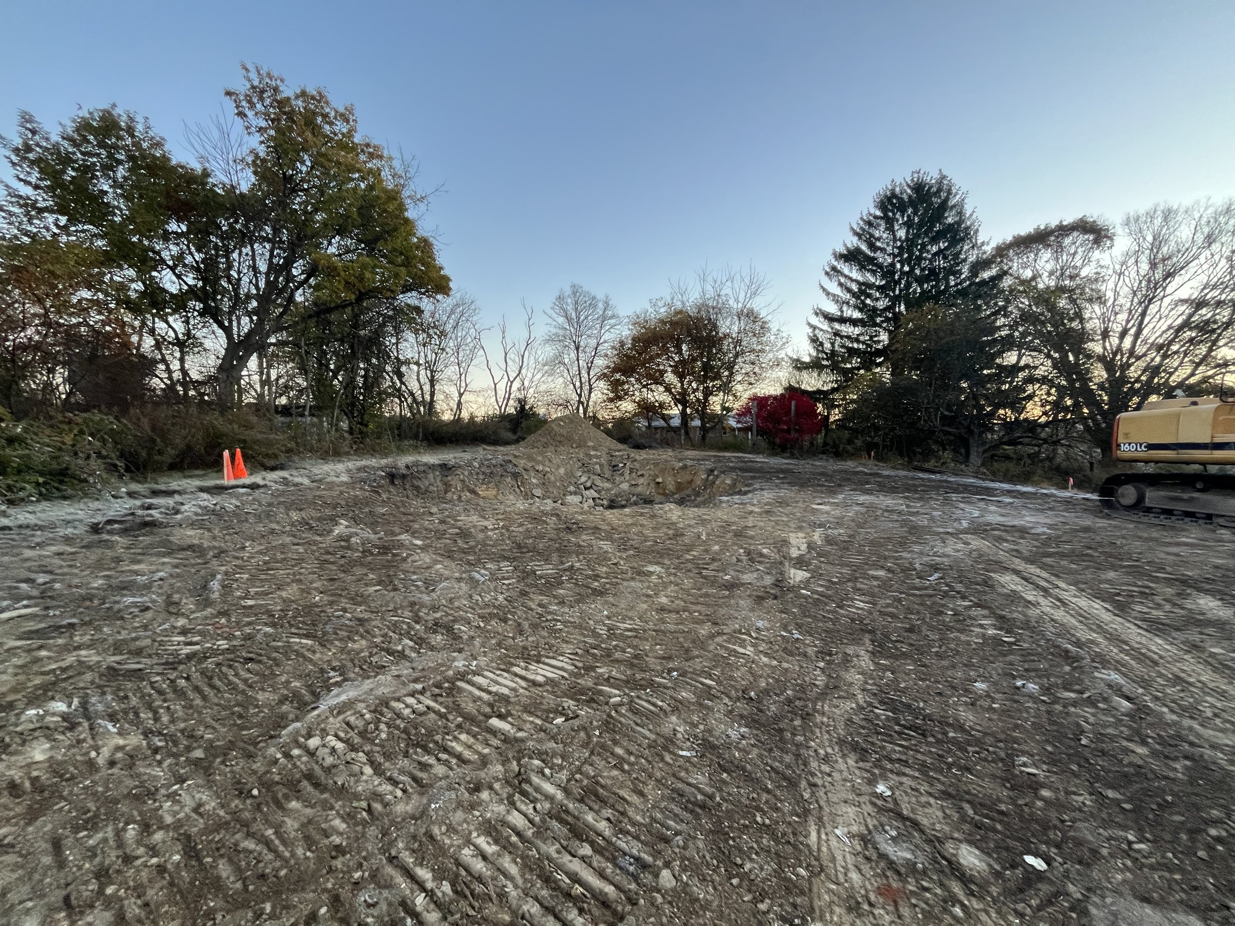 A construction site with dirt and tire tracks, orange safety cones, and an excavator with a yellow arm, surrounded by trees.
